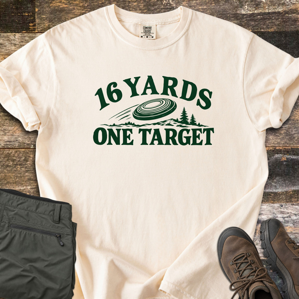 16 Yards One Target Comfort Colors T-shirt in Ivory, laid flat. Dark green print of a clay target flying over a hilly treeline with pine trees, between the arched words 16 Yards and One Target. Olive cargo pants and brown hiking boots lie beside it on weathered wood planks.