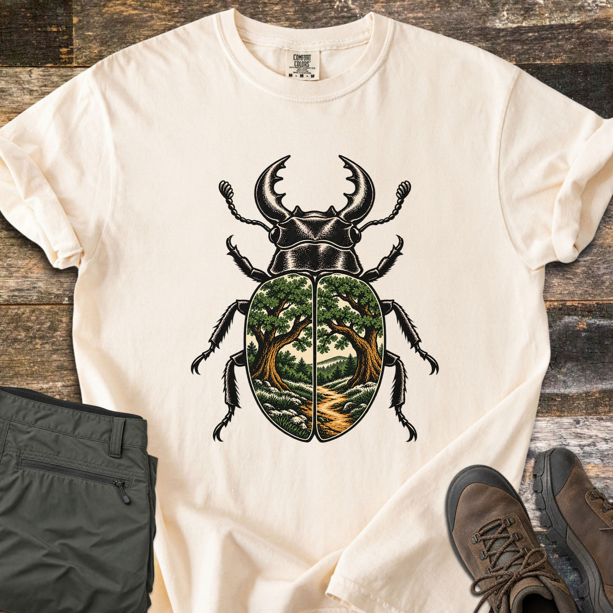 Beetle Forest Path Comfort Colors T-shirt in Ivory, laid flat. A black stag beetle whose wing covers show a forest scene of two oak trees framing a dirt path toward distant hills. Folded olive hiking pants and brown hiking boots sit beside it on weathered wood planks.
