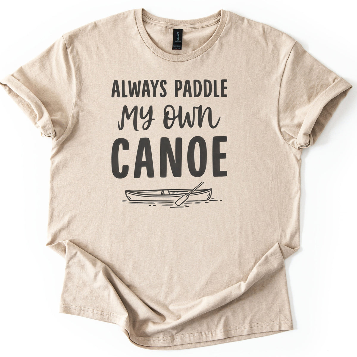 Paddle My Own Canoe Comfort Colors T-shirt in Sand, laid flat. Dark charcoal text reads Always Paddle My Own Canoe above a line drawing of a canoe with a paddle on rippling water. The shirt has rolled sleeves and lies on a white surface.