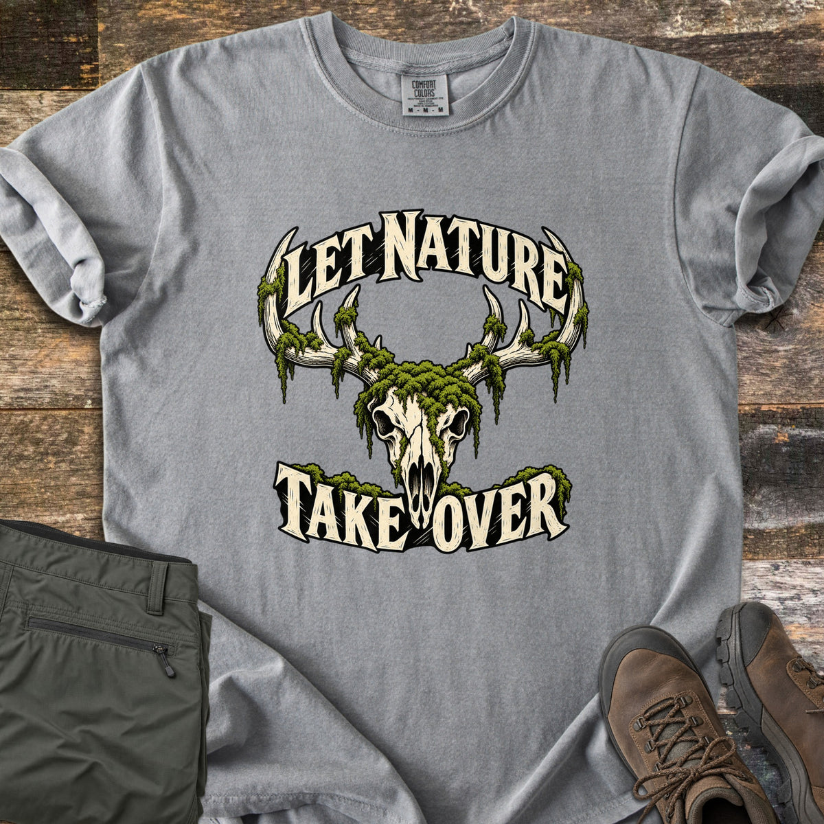 Let Nature Take Over Comfort Colors T-shirt in Grey, laid flat. A moss-covered deer skull with antlers sits between the words Let Nature Take Over in cream lettering. Folded olive hiking pants and brown hiking boots lie beside it on weathered wood planks.