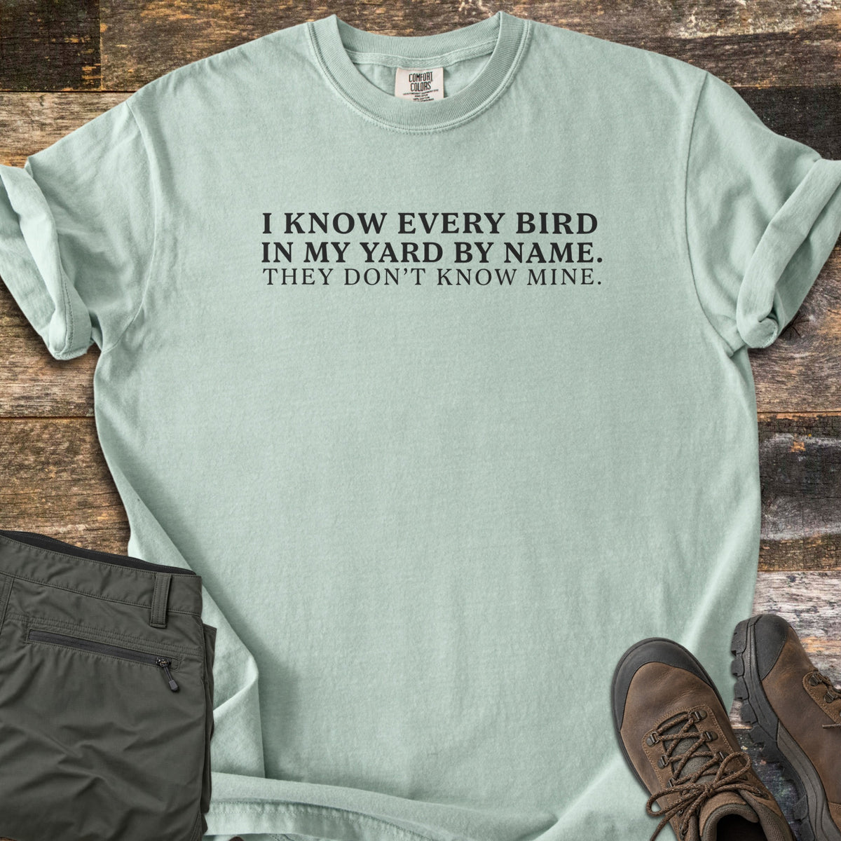 I Know Every Bird in My Yard by Name Comfort Colors T-shirt in Bay, laid flat. Black serif text reads I KNOW EVERY BIRD IN MY YARD BY NAME. THEY DON'T KNOW MINE. The sleeves are cuffed, with folded olive hiking pants and brown hiking boots on weathered wood planks.