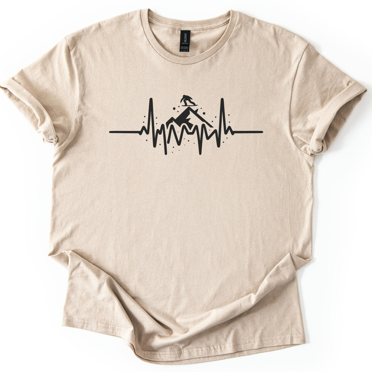 Snow Boarder Heartbeat Comfort Colors T-shirt in Sand, laid flat. A black heartbeat line rises into a mountain peak with a snowboarder silhouette riding on top, surrounded by snow dots. No words. The shirt has rolled sleeves and a rumpled hem on a white background.