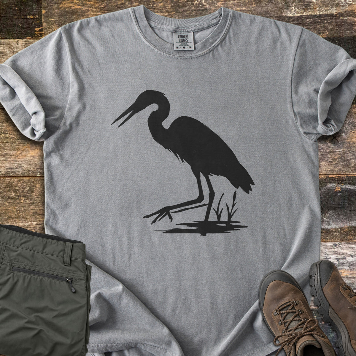 Great Blue Heron Comfort Colors T-shirt in Grey, laid flat. A black silhouette of a heron wading with its beak open, one foot raised, beside cattails over a patch of water. Sleeves rolled, on weathered wood with olive hiking pants and brown hiking boots.