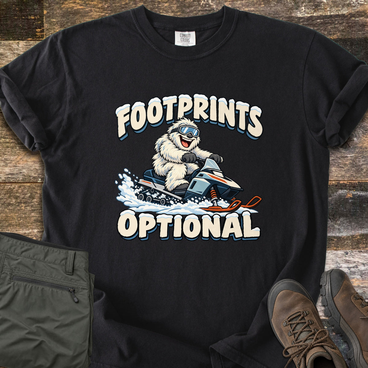 Footprints Optional Comfort Colors T-shirt in Black, laid flat. A grinning white yeti in ski goggles rides a snowmobile through spraying snow between the snow-capped words Footprints Optional. It lies on weathered wood beside folded olive hiking pants and a pair of brown boots.
