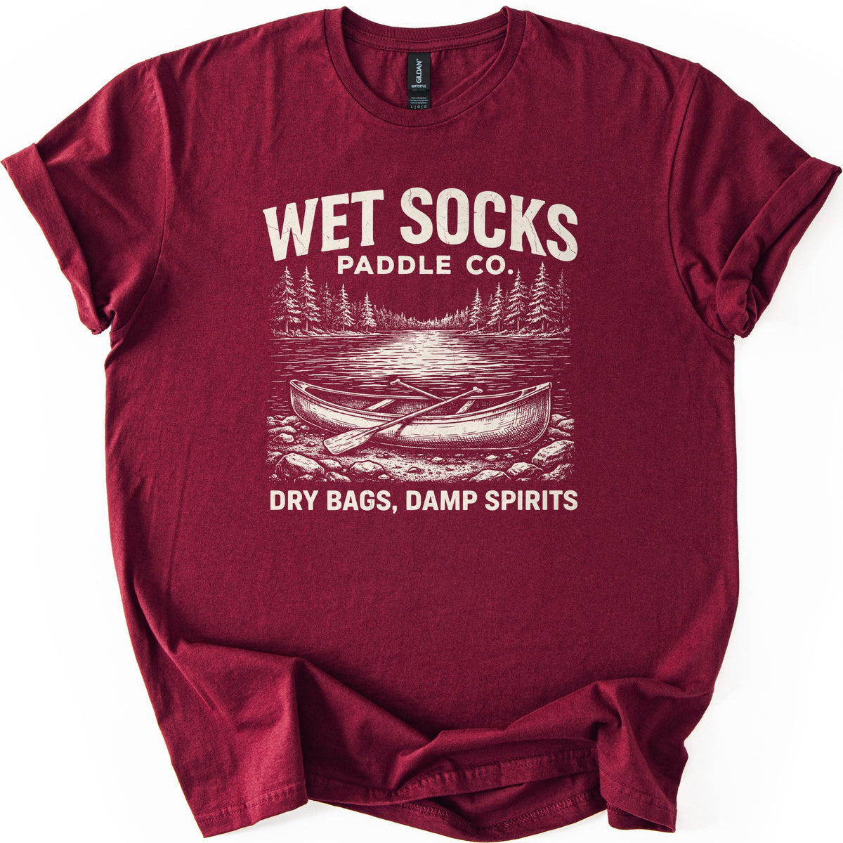Wet Socks Paddle Co Comfort Colors T-shirt in Maroon, laid flat. A cream etched drawing of a canoe with crossed paddles on a rocky shore, with a lake and pine trees behind, under the words Wet Socks Paddle Co. and above Dry Bags, Damp Spirits. White background.