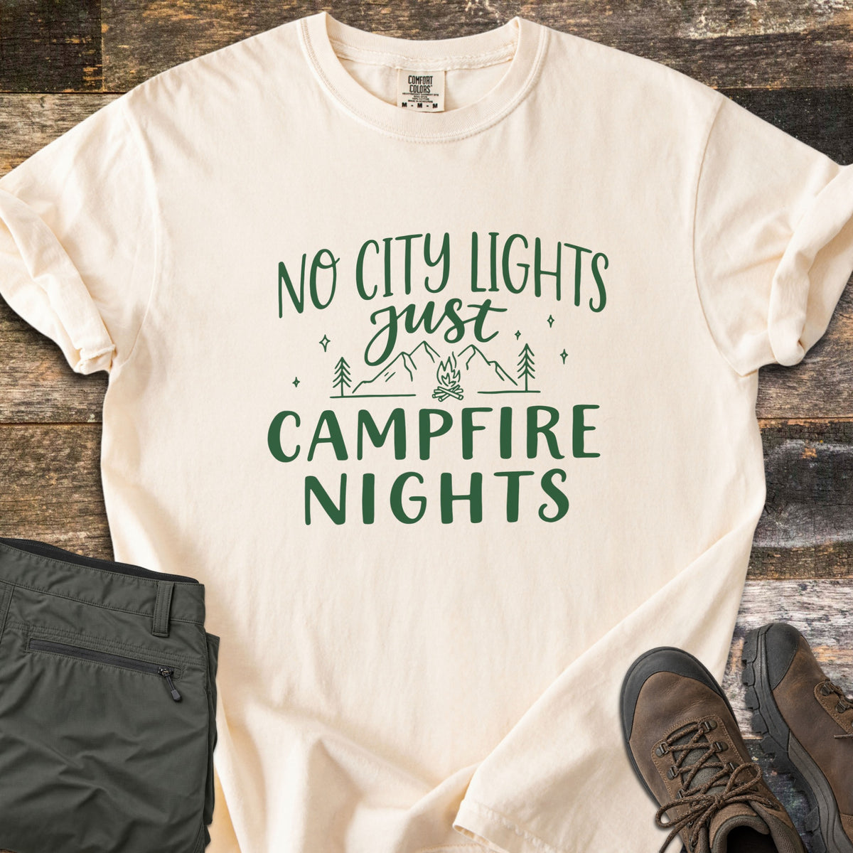 Campfire Nights Comfort Colors T-shirt in Ivory, laid flat. Dark green line art of mountains, pine trees, stars and a campfire sits between the words No City Lights just Campfire Nights. On rustic wood planks with folded olive hiking pants and brown boots.