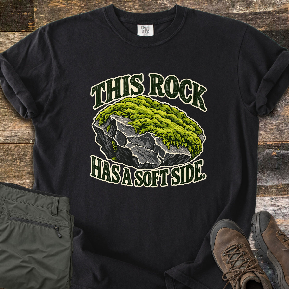 This Rock Has a Soft Side Comfort Colors T-shirt in Black, laid flat. An illustrated gray boulder topped with thick green moss sits between the words This Rock above and Has a Soft Side. below. Olive hiking pants and brown hiking boots lie beside it on weathered wood planks.