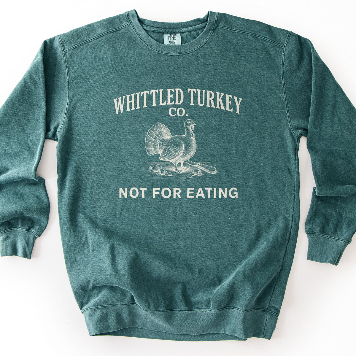 Whittled Turkey Co Comfort Colors crewneck sweatshirt in Blue Spruce, laid flat. Cream print of a vintage-style engraved turkey standing on a wood carving with curled shavings and a whittling knife, under the words Whittled Turkey Co. and above Not For Eating. Shown on a white background.