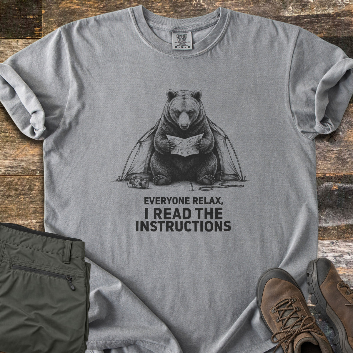 Everyone Relax I Read The Instructions Comfort Colors T-shirt in Grey, laid flat. A black sketch of a bear reading instructions in front of a tent, with a tipped lantern and rope, above the words Everyone relax, I read the instructions. Olive cargo pants and brown hiking boots sit on dark wood.
