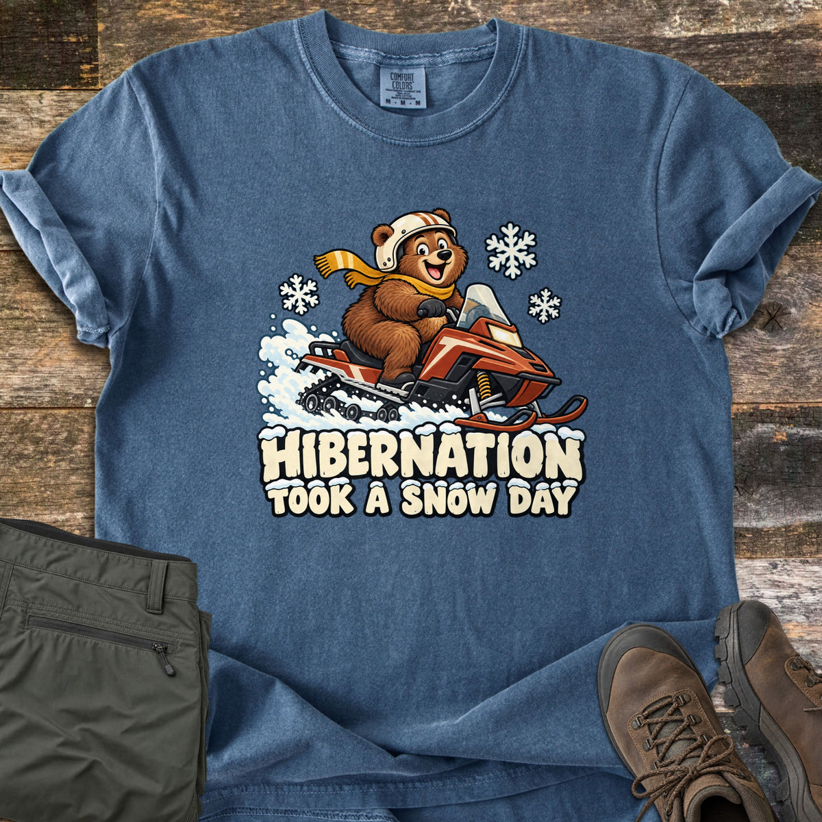 Hibernation Took a Snow Day Comfort Colors T-shirt in Denim, laid flat. A cartoon bear in a white helmet and yellow scarf rides a red snowmobile through snow among snowflakes, above the words Hibernation Took a Snow Day. Olive pants and brown boots sit beside it on weathered wood.
