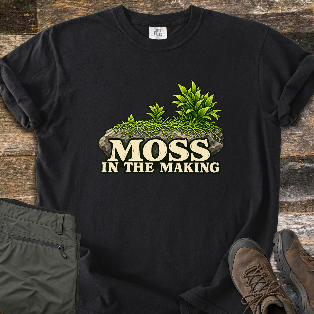 Moss in the Making Comfort Colors T-shirt in Black, laid flat. A mossy gray rock sprouting green leafy plants sits above the words Moss in the Making. Olive hiking pants and brown hiking boots lie beside it on weathered wood planks.