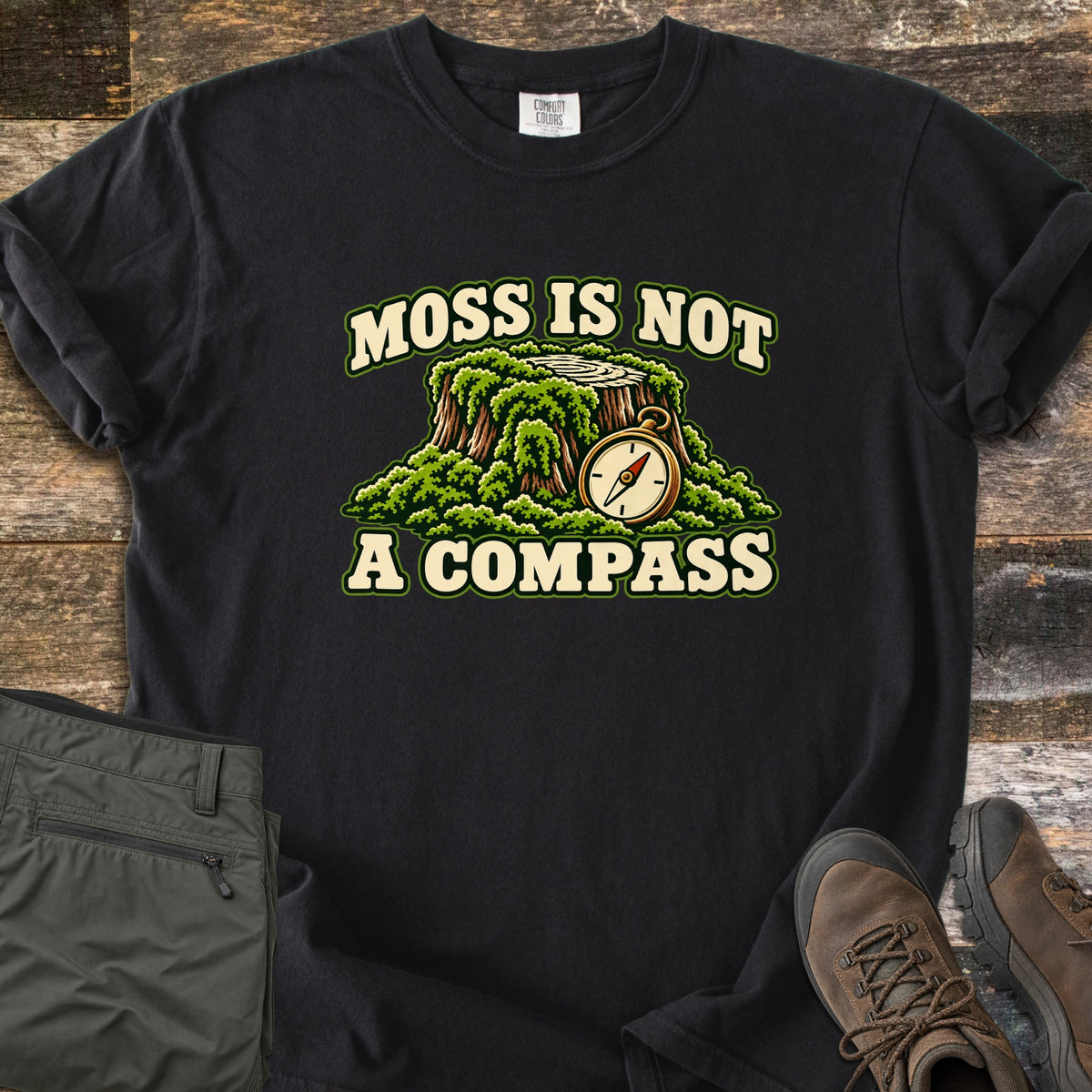 Moss Is Not a Compass Comfort Colors T-shirt in Black, laid flat. A mossy tree stump with a brass compass leaning on it is framed by the words MOSS IS NOT and A COMPASS. Folded olive hiking pants and brown hiking boots sit on rustic wood planks.