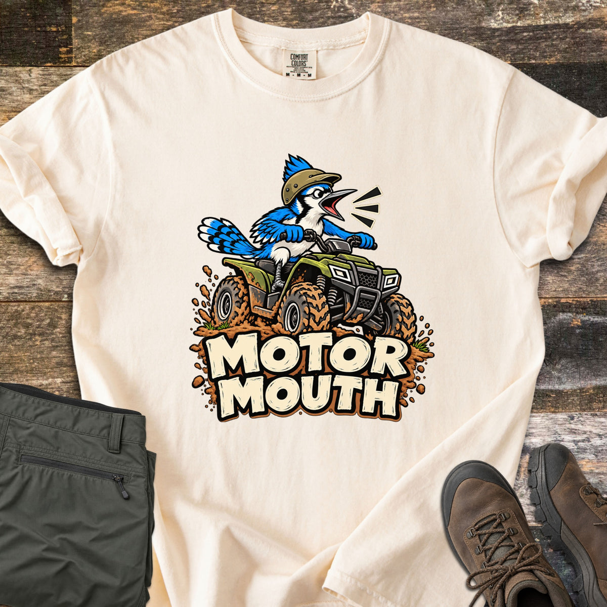 Motor Mouth Comfort Colors T-shirt in Ivory, laid flat. A cartoon blue jay in a tan helmet yells while riding a green four-wheeler through mud above the words Motor Mouth. Folded olive pants and brown hiking boots sit beside the shirt on rustic wood.