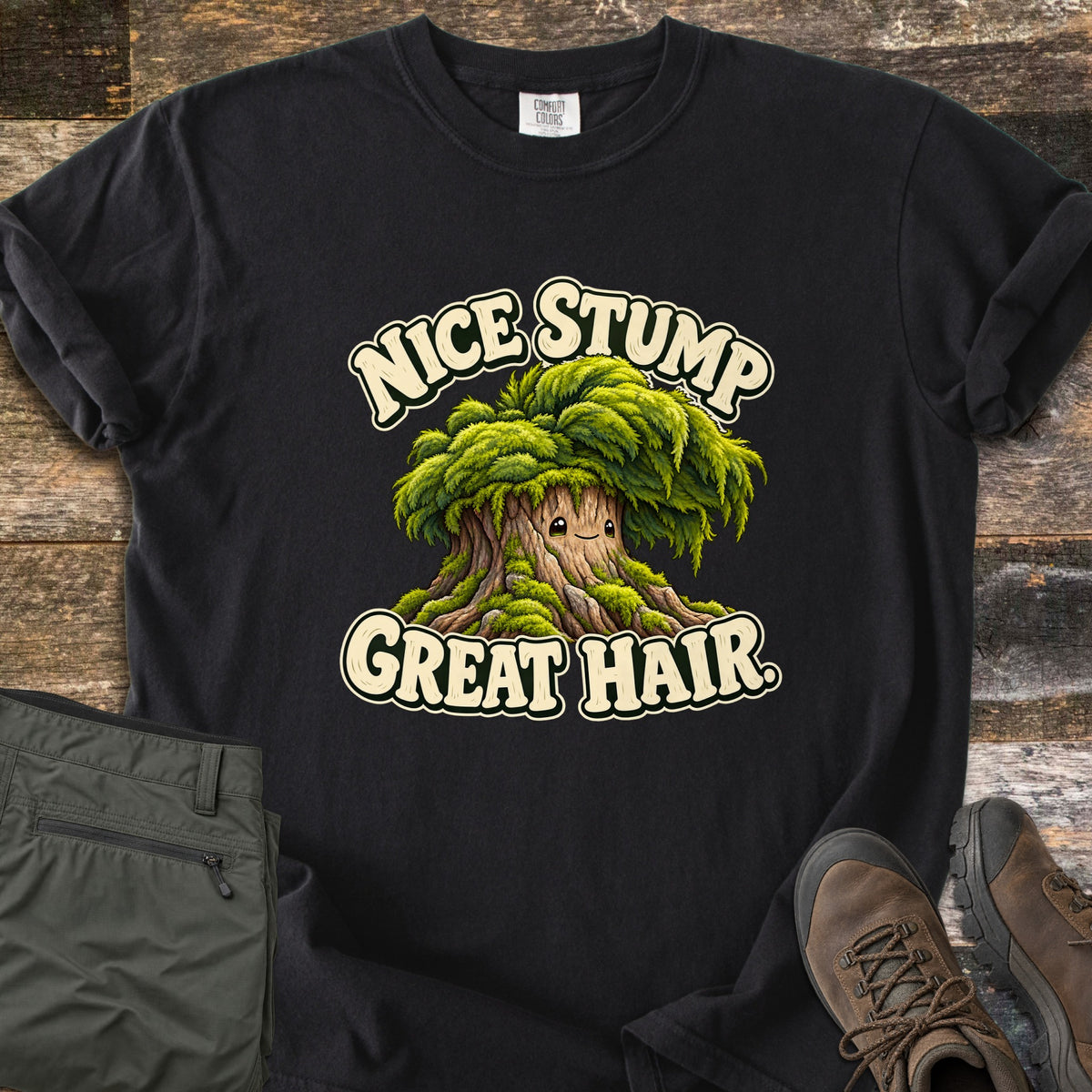Nice Stump Great Hair Comfort Colors T-shirt in Black, laid flat. A smiling cartoon tree stump with a shaggy mop of green moss hair sits between the words Nice Stump Great Hair. Sleeves rolled, on weathered wood with folded olive hiking pants and brown hiking boots.