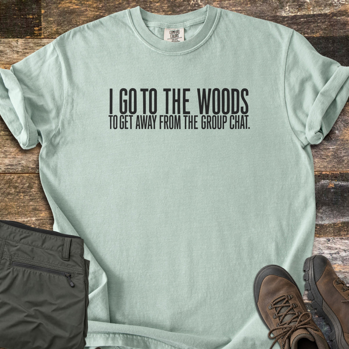 Away From The Group Chat Comfort Colors T-shirt in Bay, laid flat. Black block letters read I GO TO THE WOODS above TO GET AWAY FROM THE GROUP CHAT. The shirt has rolled sleeves and lies on weathered wood boards beside folded olive green pants and brown hiking boots.