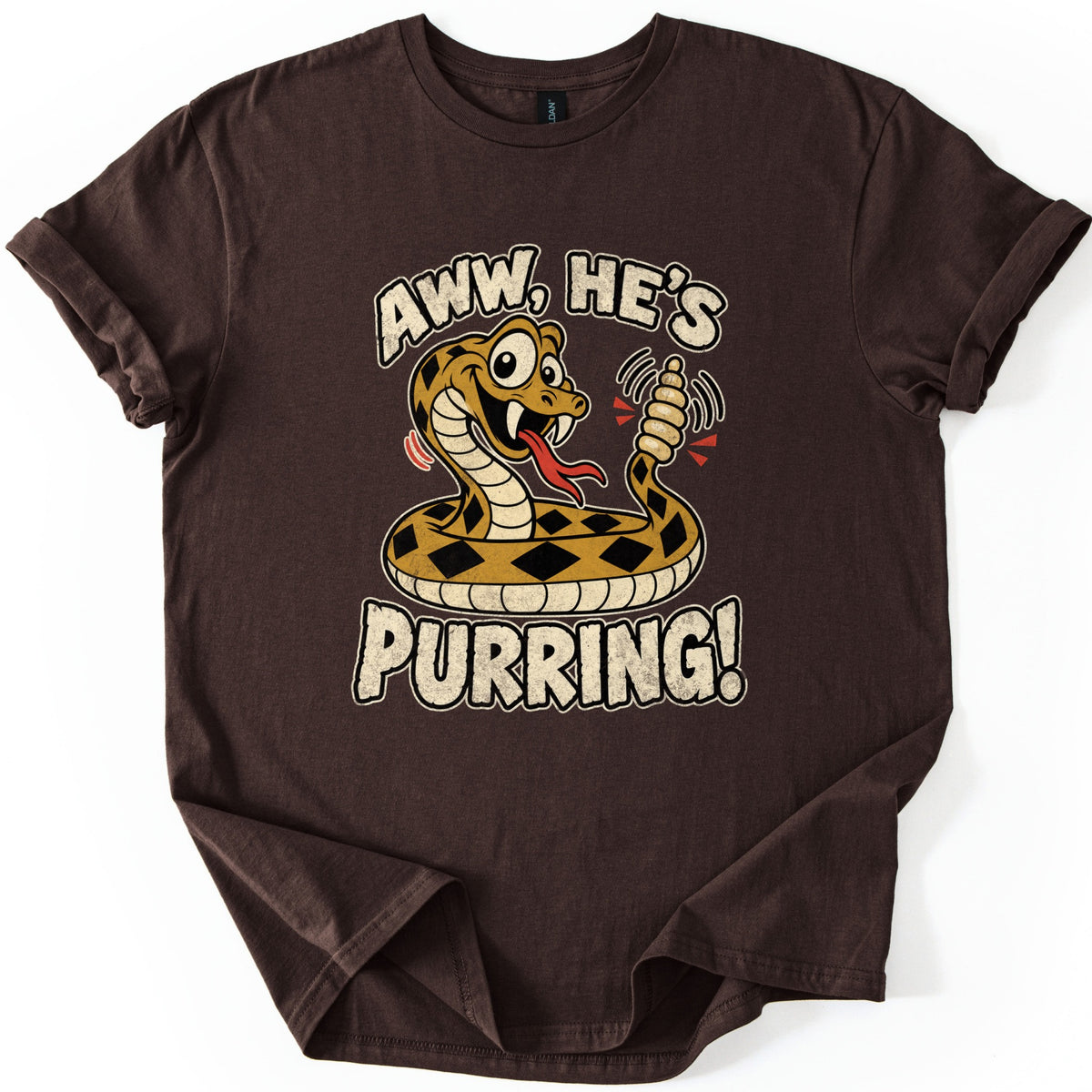 Aww He's Purring Comfort Colors T-shirt in Dark Chocolate, laid flat. A cartoon rattlesnake with diamond-patterned scales grins and shakes its rattle between the words Aww, He's Purring! in worn cream letters. The shirt lies on a white background.
