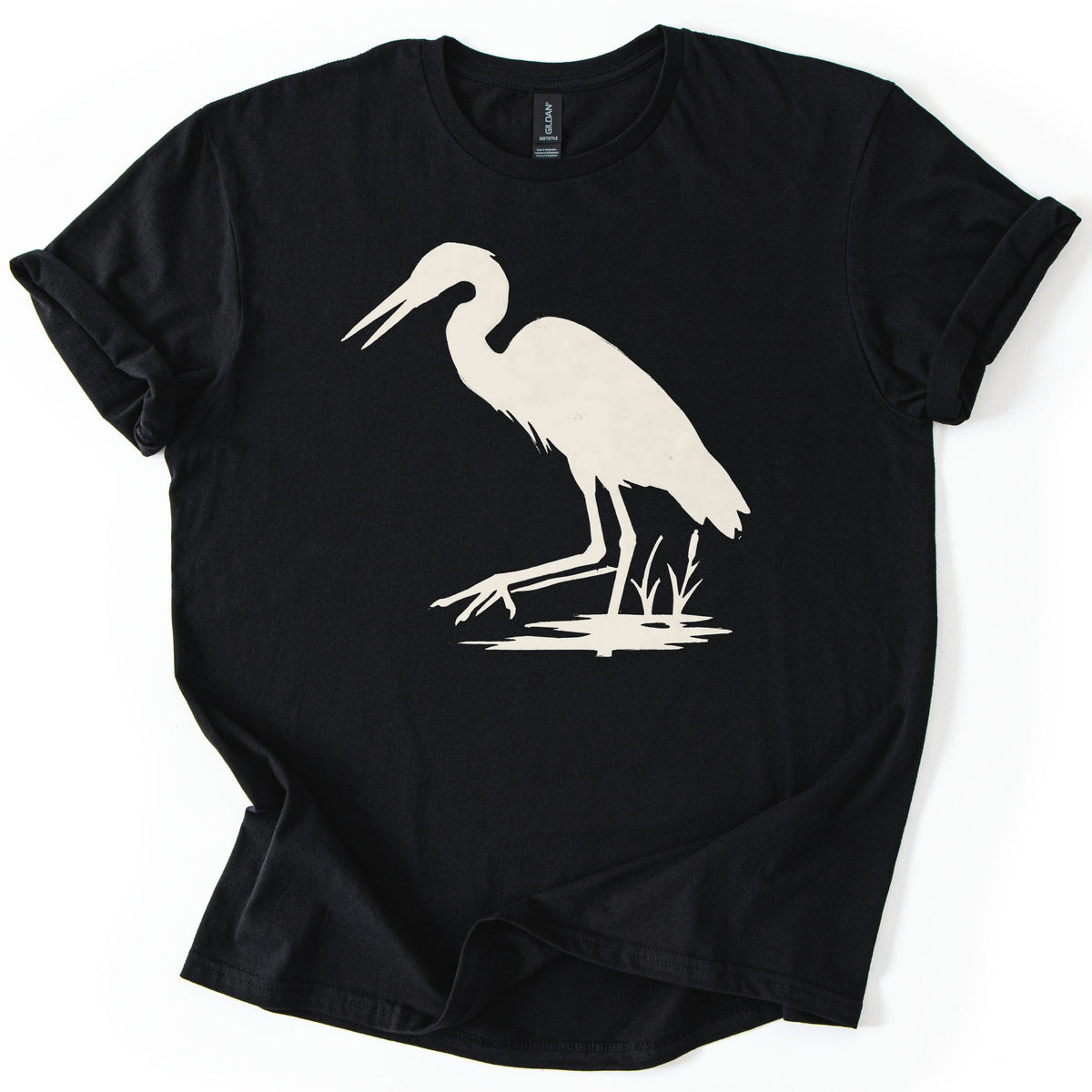Great Blue Heron Comfort Colors T-shirt in Black, laid flat. A cream silhouette of a heron wading in shallow water beside cattails, beak open and one foot reaching forward. The shirt lies on a white surface with cuffed sleeves.