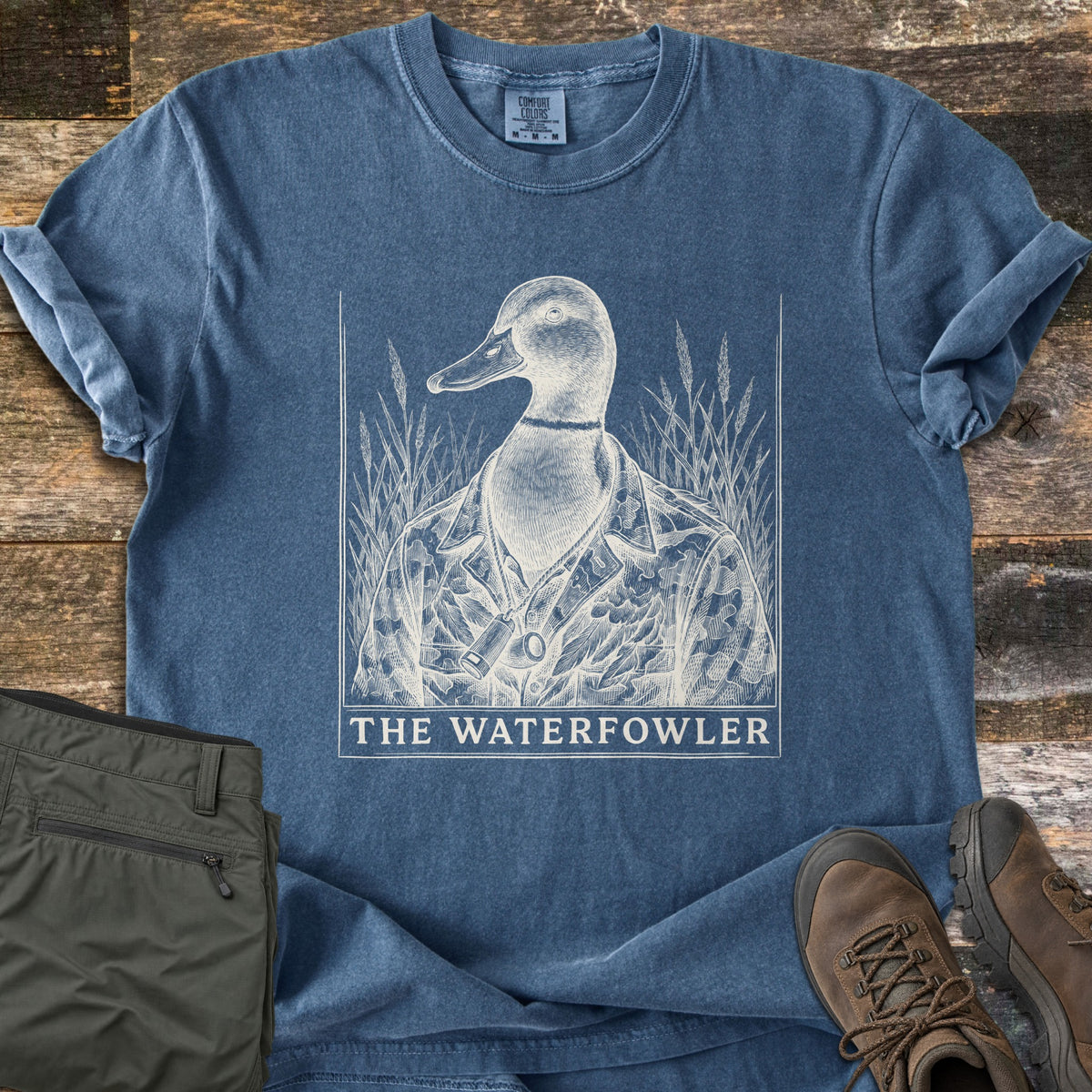 The Waterfowler Comfort Colors T-shirt in Denim, laid flat. A cream sketch-style drawing shows a mallard duck in a camo jacket with a duck call on a lanyard, framed by marsh grass above the words The Waterfowler. Olive pants and brown boots sit on weathered wood.
