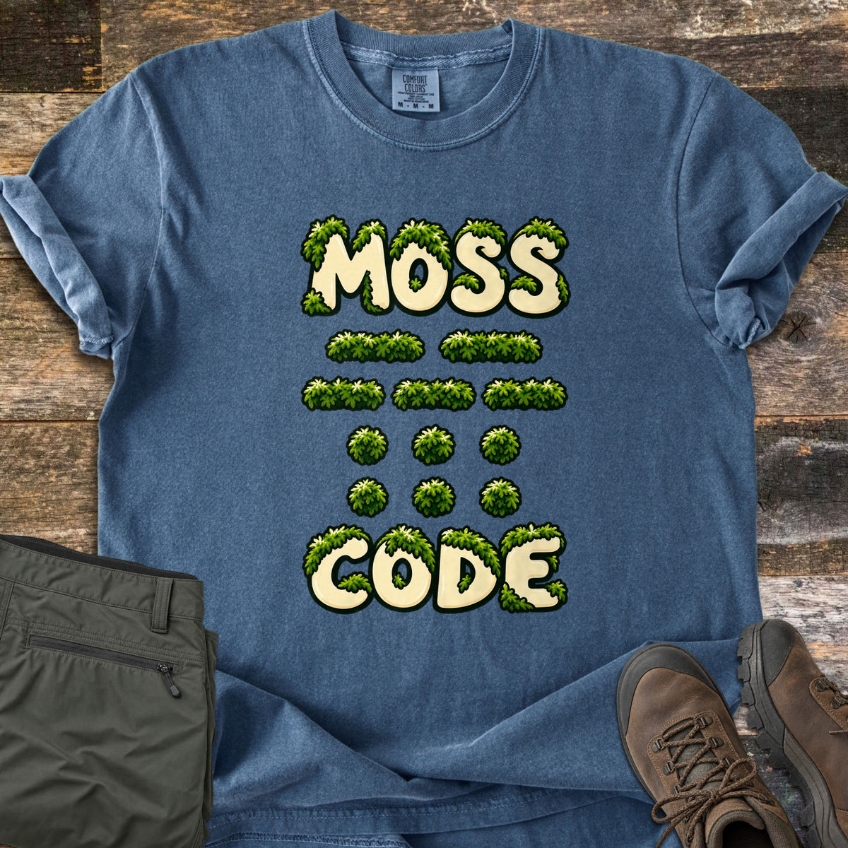 Moss Code Comfort Colors T-shirt in Denim, laid flat. Cream letters draped in green moss spell MOSS, then rows of moss clump dashes and dots in Morse code, then CODE. Sleeves rolled, on weathered wood planks beside olive pants and brown hiking boots.