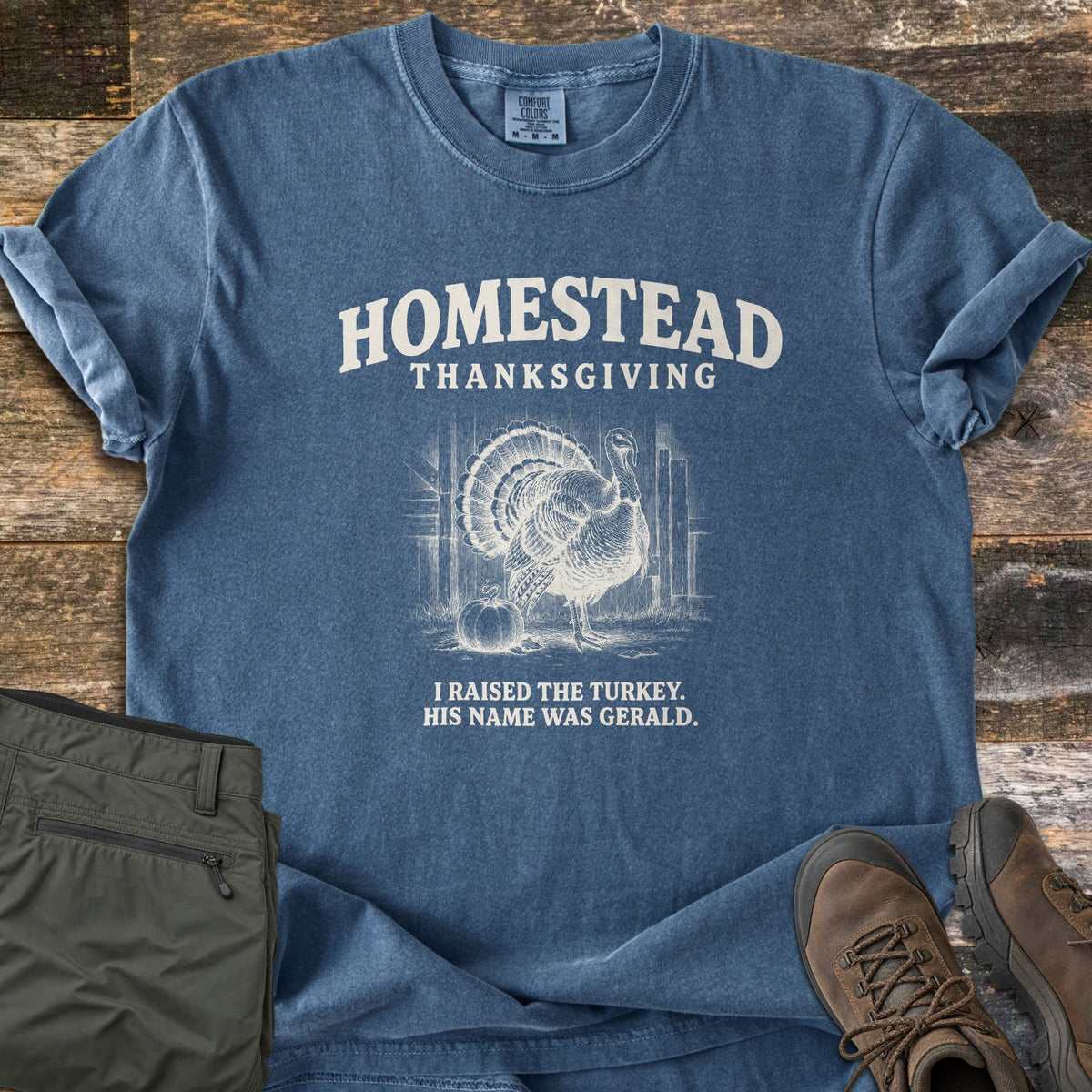 Homestead Thanksgiving Comfort Colors T-shirt in Denim, laid flat. Homestead Thanksgiving arches over a white sketch of a turkey beside a pumpkin in a barn, above the words I raised the turkey. His name was Gerald. On weathered wood with olive pants and brown hiking boots.