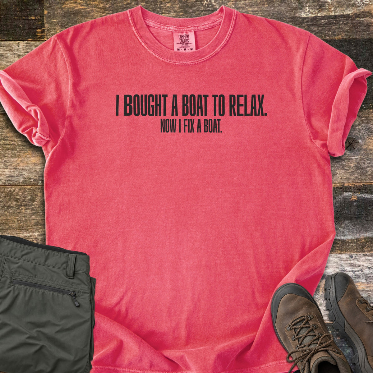 Boat To Relax Comfort Colors T-shirt in Watermelon, laid flat. Black block letters read I BOUGHT A BOAT TO RELAX. with NOW I FIX A BOAT. in smaller type below. Sleeves are cuffed, with folded olive cargo pants and brown hiking boots on weathered wood planks.
