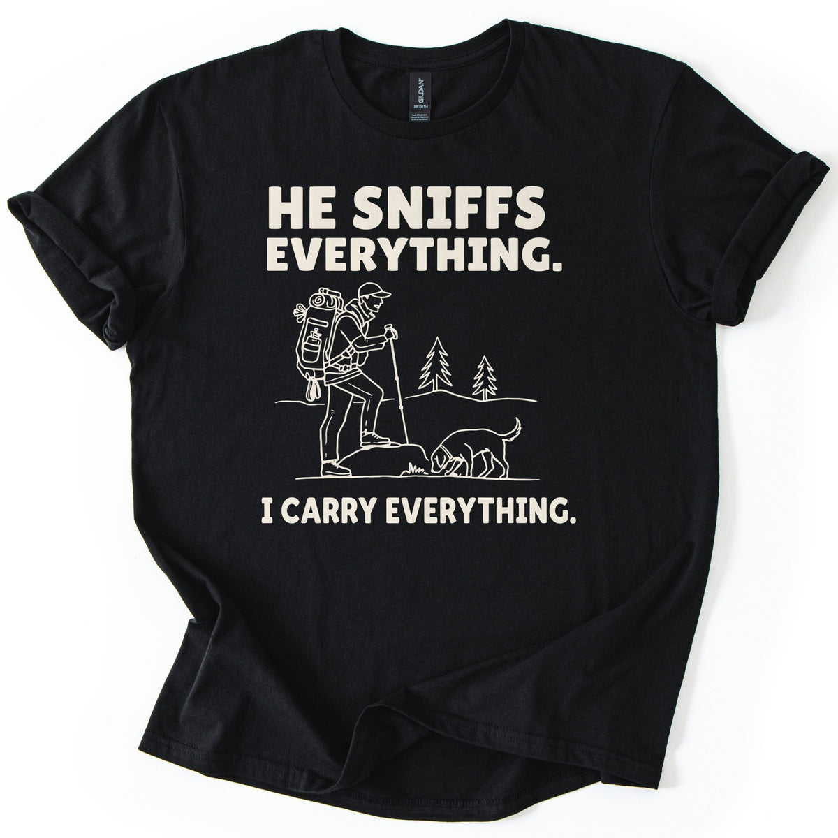 Trail Dog Pack Mule Comfort Colors T-shirt in Black, laid flat. Cream line art of a hiker with a loaded backpack and trekking pole stepping onto a rock as a dog sniffs the ground, with pine trees behind, between the words He sniffs everything. and I carry everything. Sleeves rolled, on white.