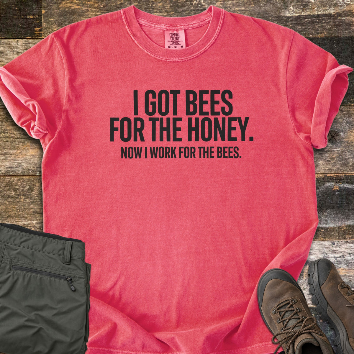 Work For The Bees Comfort Colors T-shirt in Watermelon, laid flat. Black block letters read I GOT BEES FOR THE HONEY. NOW I WORK FOR THE BEES. The shirt lies with cuffed sleeves on weathered wood planks beside folded olive green pants and brown hiking boots.