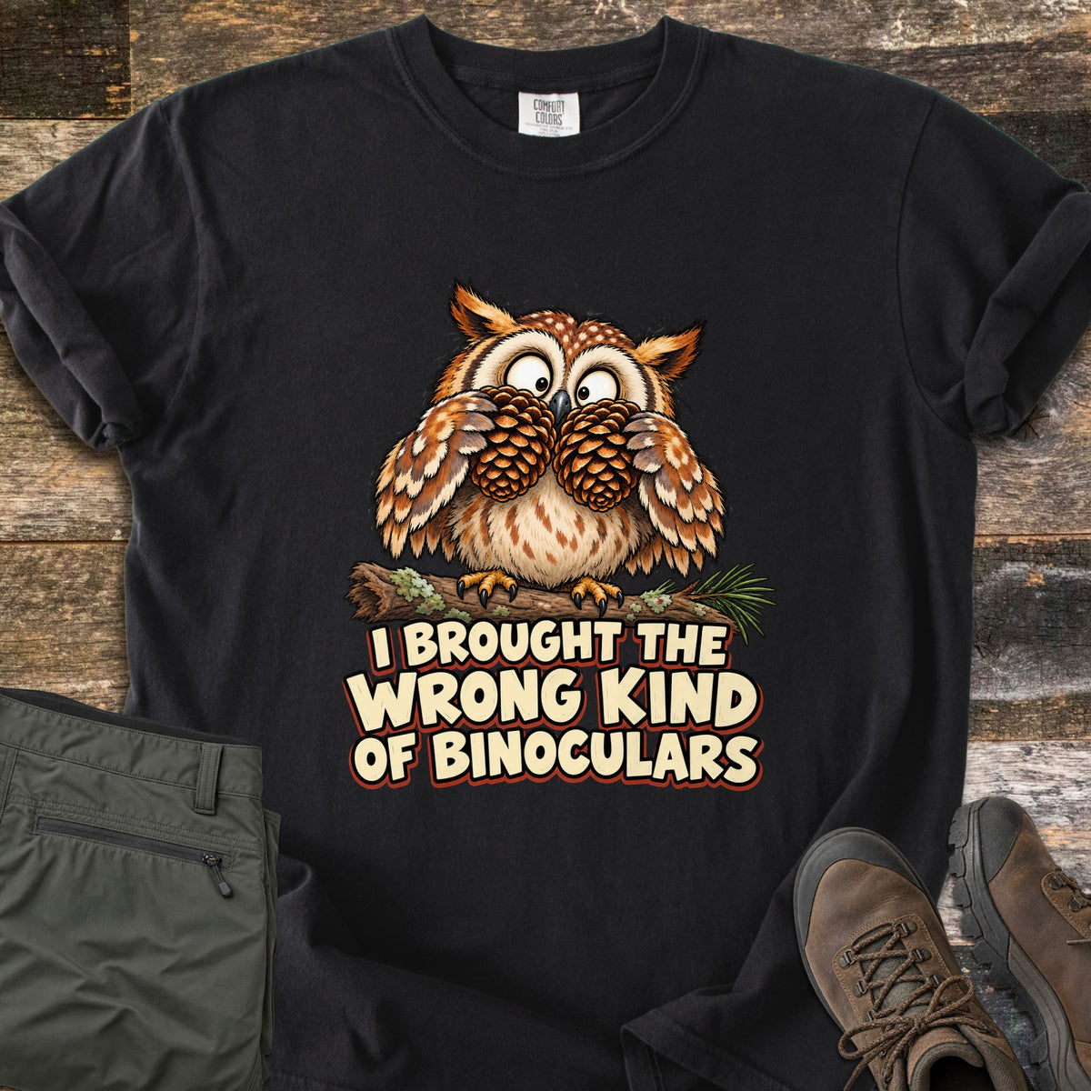 I Brought The Wrong Kind Of Binoculars Comfort Colors T-shirt in Black, laid flat. A wide-eyed cartoon owl on a mossy branch holds two pinecones to its eyes like binoculars above the words I Brought The Wrong Kind Of Binoculars. Olive hiking pants and brown hiking boots lie beside it on weathered wood.