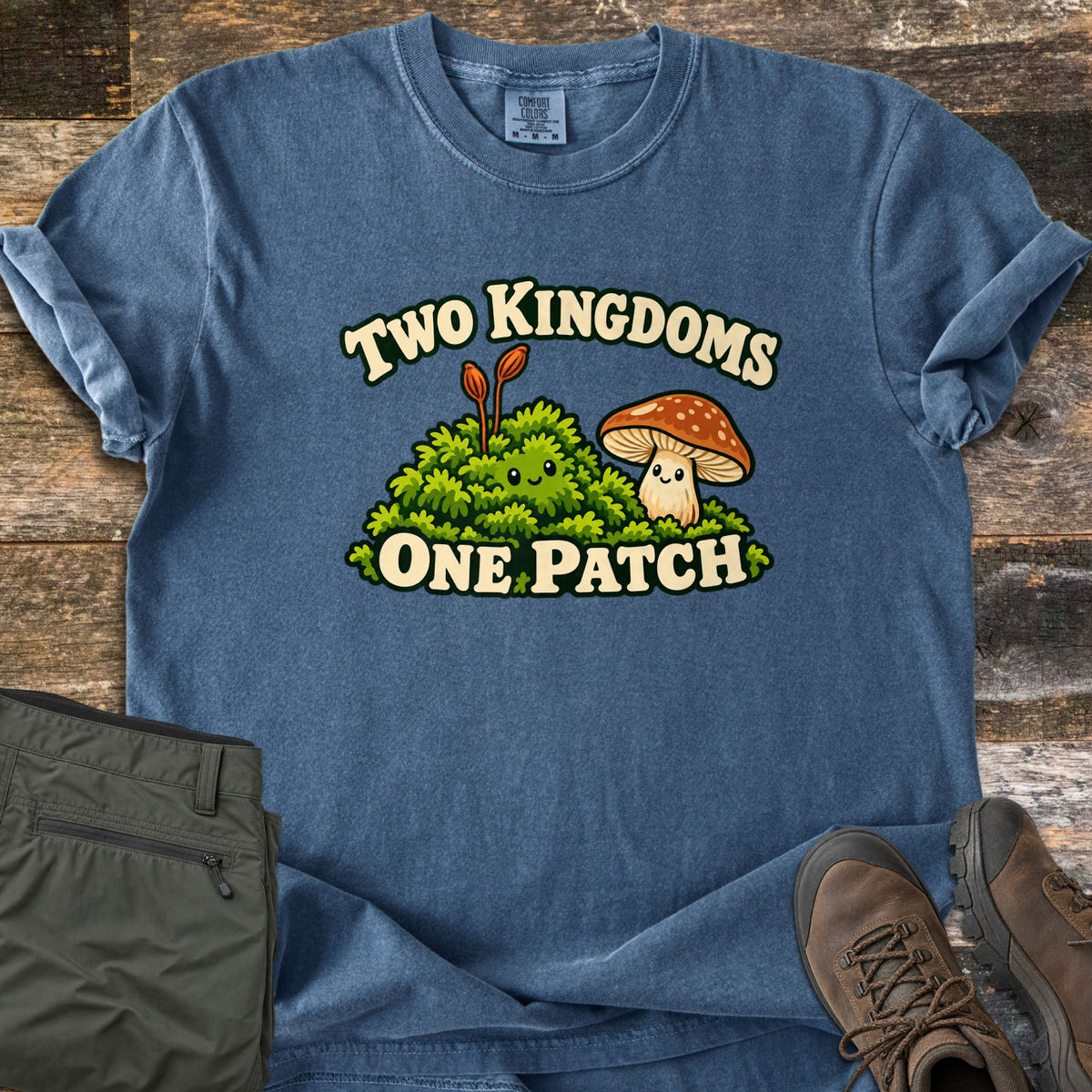Two Kingdoms One Patch Comfort Colors T-shirt in Denim, laid flat. A smiling green moss clump with two sprouts sits beside a smiling orange-capped mushroom, between the words Two Kingdoms One Patch. Olive hiking pants and brown hiking boots lie beside it on dark wood planks.