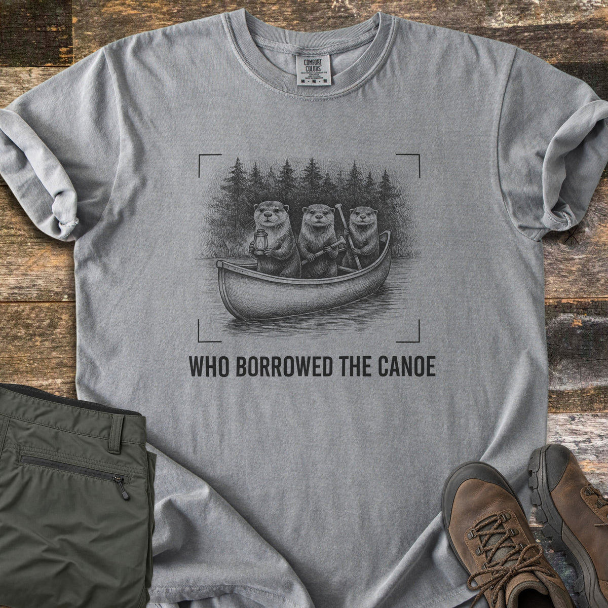 Who Borrowed The Canoe Comfort Colors T-shirt in Grey, laid flat. A black sketch of three otters in a canoe before pine trees, one holding a lantern and two holding paddles, framed by camera corner marks above the words Who Borrowed The Canoe. Olive pants and brown boots nearby.