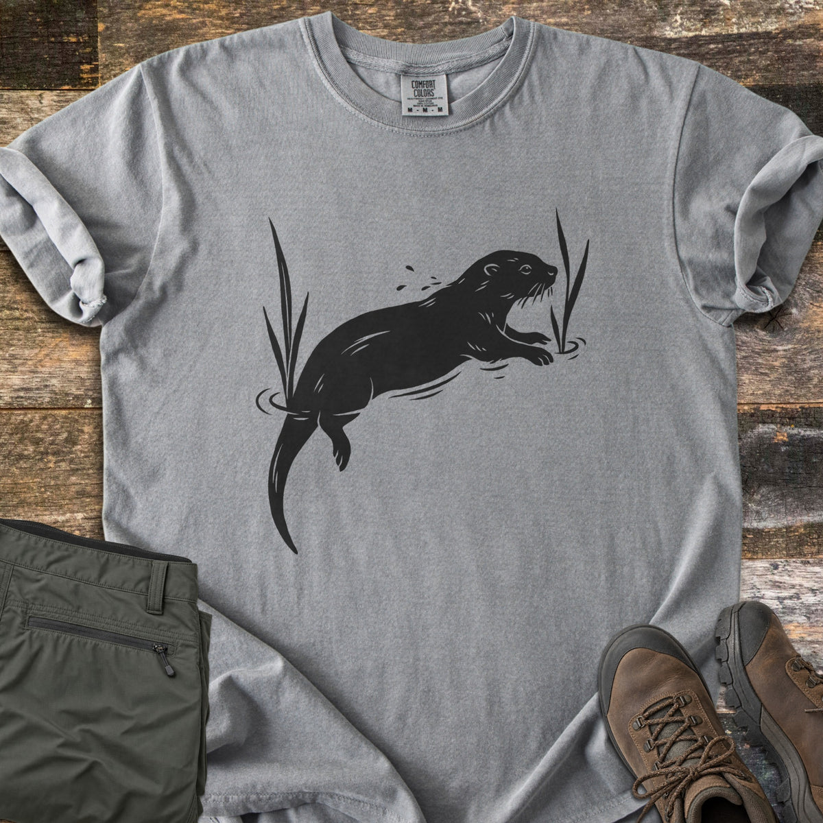 Swimming Otter Comfort Colors T-shirt in Grey, laid flat. A black print of an otter swimming between tall reeds, with ripples and splashing droplets. Sleeves are rolled; olive hiking pants and brown hiking boots sit on weathered wood planks.