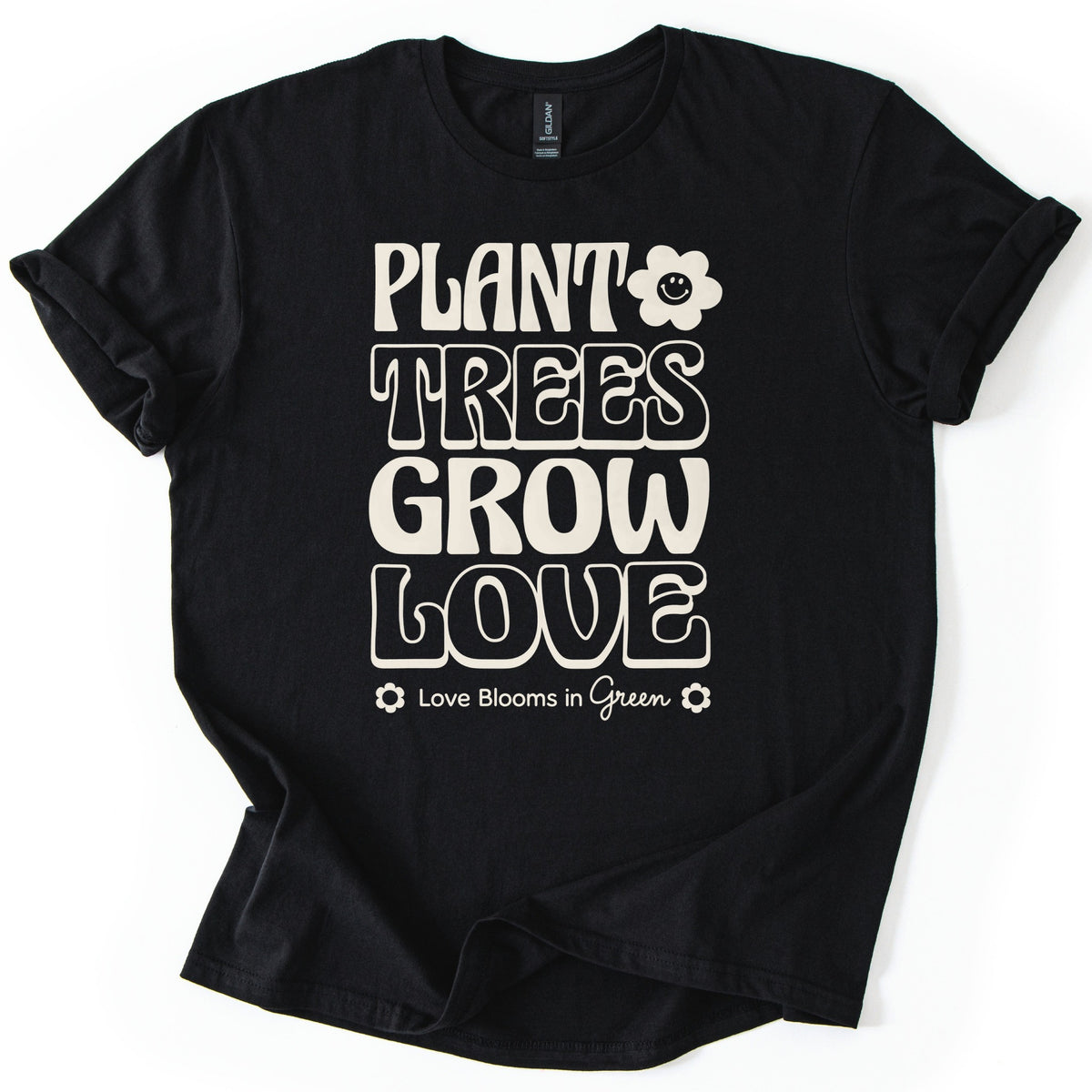Plant Trees Grow Love Comfort Colors T-shirt in Black, laid flat. Cream retro bubble letters read Plant Trees Grow Love, with a smiling daisy beside Plant and Love Blooms in Green between two small flowers below. Sleeves are rolled on a white background.