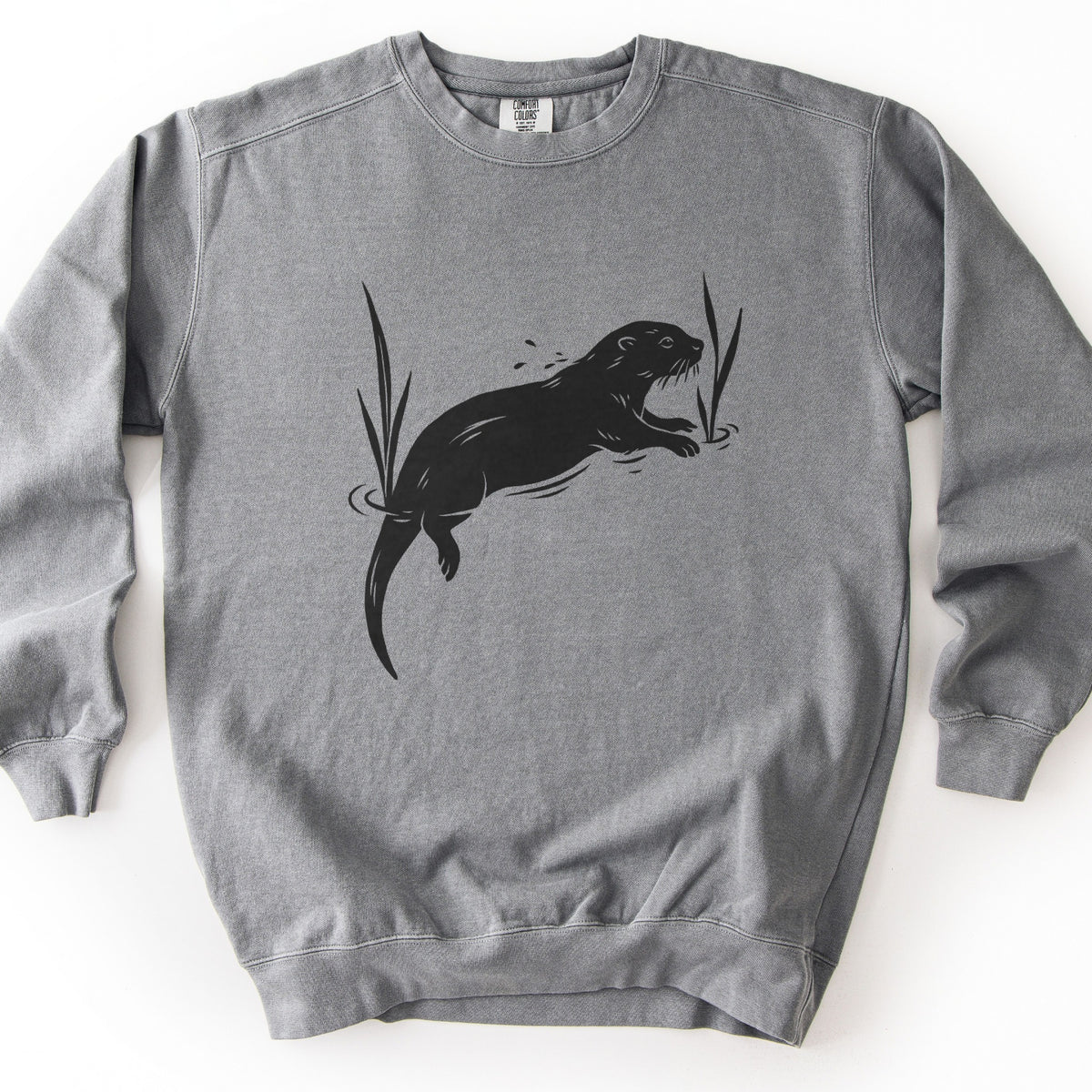 Swimming Otter Comfort Colors crewneck sweatshirt in Grey, laid flat. A black block-print style otter swims with its head raised, splashing droplets, between two tall reeds with ripples around them. No words are printed. The sweatshirt lies on a white background.