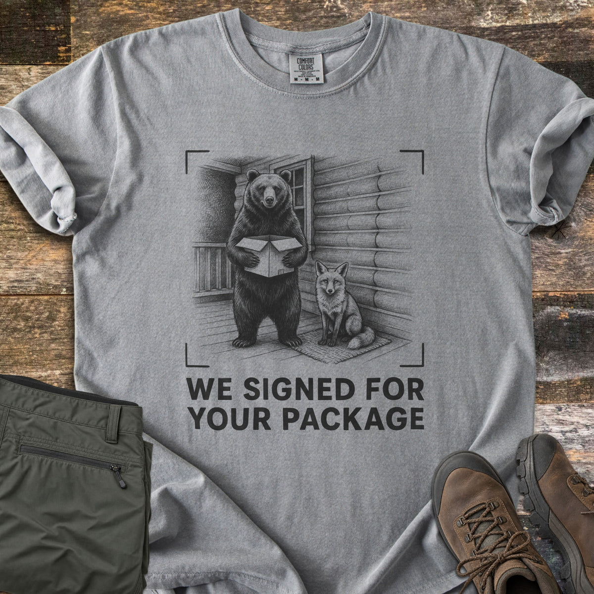 We Signed For Your Package Comfort Colors T-shirt in Grey, laid flat. A black-ink sketch shows a bear holding an open box beside a seated fox on a log cabin porch, framed by camera corner marks above the words We Signed For Your Package. Olive pants and brown hiking boots sit on wood planks.