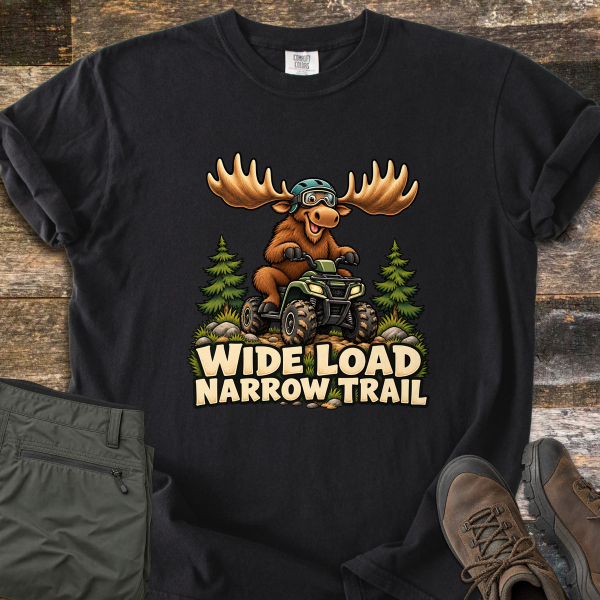 Wide Load Narrow Trail Comfort Colors T-shirt in Black, laid flat. A cartoon moose with big antlers, a helmet and goggles rides a green ATV between pine trees above the words Wide Load Narrow Trail. Folded olive pants and brown hiking boots lie beside it on weathered wood.