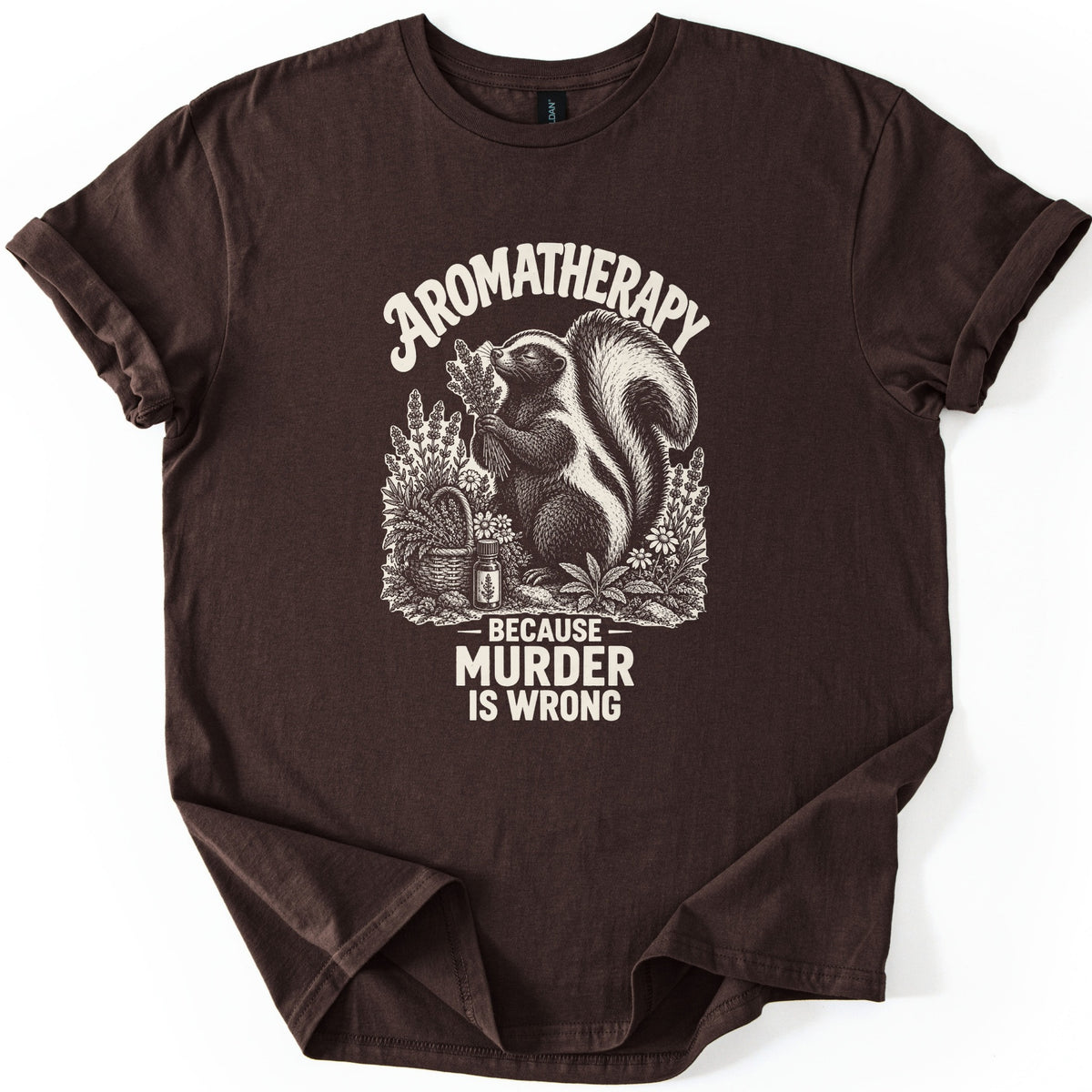 Aromatherapy Murder Skunk Comfort Colors T-shirt in Dark Chocolate, laid flat. A cream skunk sniffs a lavender bundle beside a basket, flowers and an oil bottle, under the word Aromatherapy and above Because Murder Is Wrong. Sleeves are cuffed on a white background.