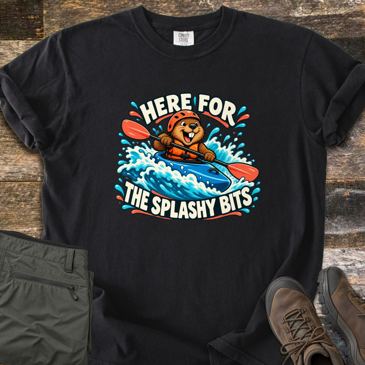 Splashy Bits Comfort Colors T-shirt in Black, laid flat. A cartoon beaver in an orange helmet and life vest paddles a blue kayak through whitewater, framed by the words Here For The Splashy Bits. Olive pants and brown hiking boots lie beside it on weathered wood.