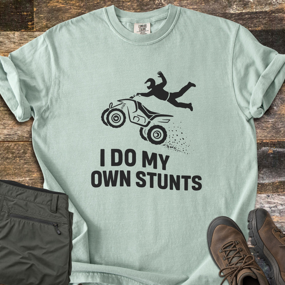 ATV Stunts Comfort Colors T-shirt in Bay, laid flat. A black silhouette of a helmeted rider flying off a wheelie-popping ATV kicking up dirt, above the words I Do My Own Stunts. Olive cargo pants and brown hiking boots sit beside it on weathered wood.