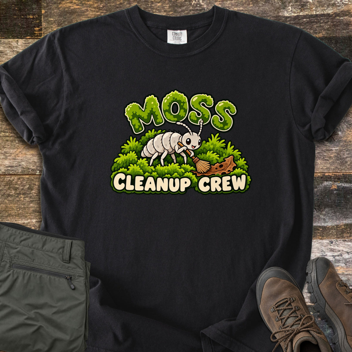 Moss Cleanup Crew Comfort Colors T-shirt in Black, laid flat. A smiling white cartoon bug sweeps a brown leaf with a broom atop green moss, below the word MOSS in mossy letters and above CLEANUP CREW. It lies on weathered wood with folded olive pants and brown hiking boots.