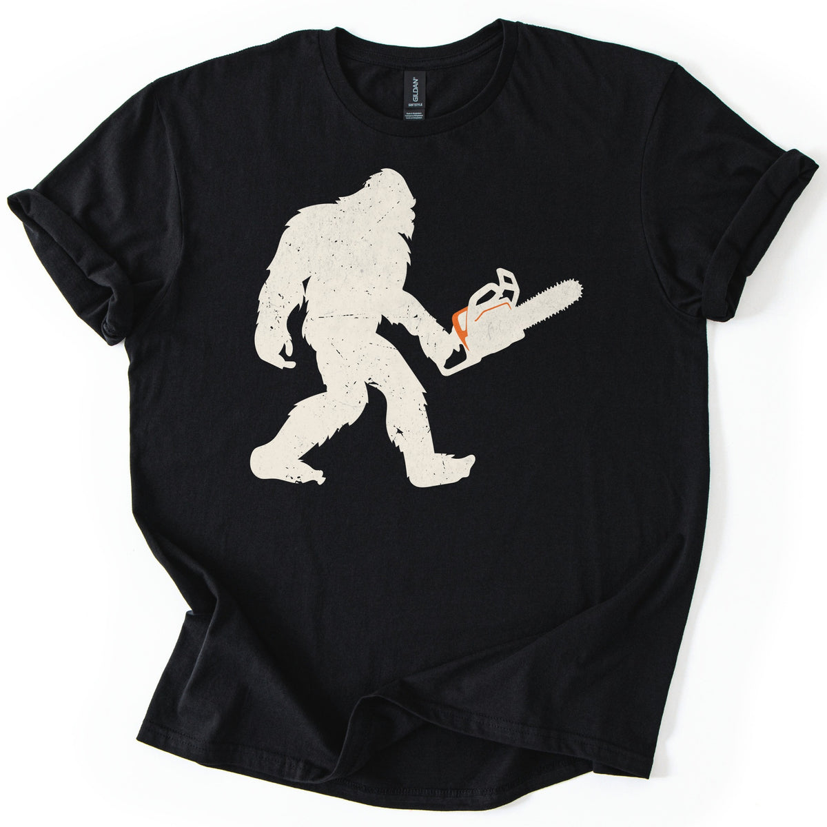 Lumber Squatch Comfort Colors T-shirt in Black, laid flat. A distressed cream silhouette of Bigfoot walking mid-stride while holding a chainsaw with an orange accent, with no words printed. The shirt has cuffed sleeves and lies on a white background.