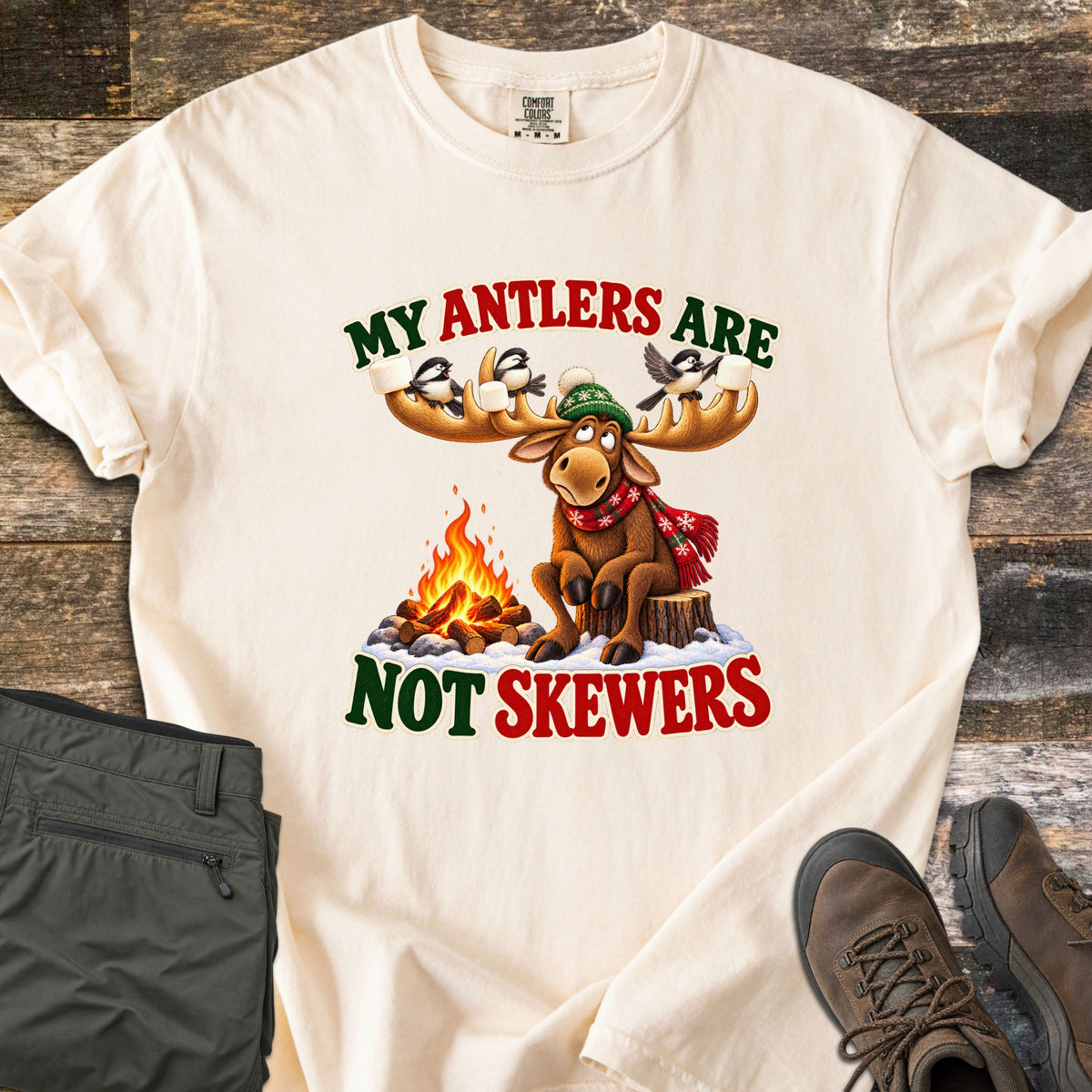 My Antlers Are Not Skewers Comfort Colors T-shirt in Ivory, laid flat. A cartoon moose in a green knit hat and red scarf sits on a stump by a campfire as chickadees on its antlers hold marshmallows, with the words My Antlers Are Not Skewers. Olive pants and brown boots lie on weathered wood.