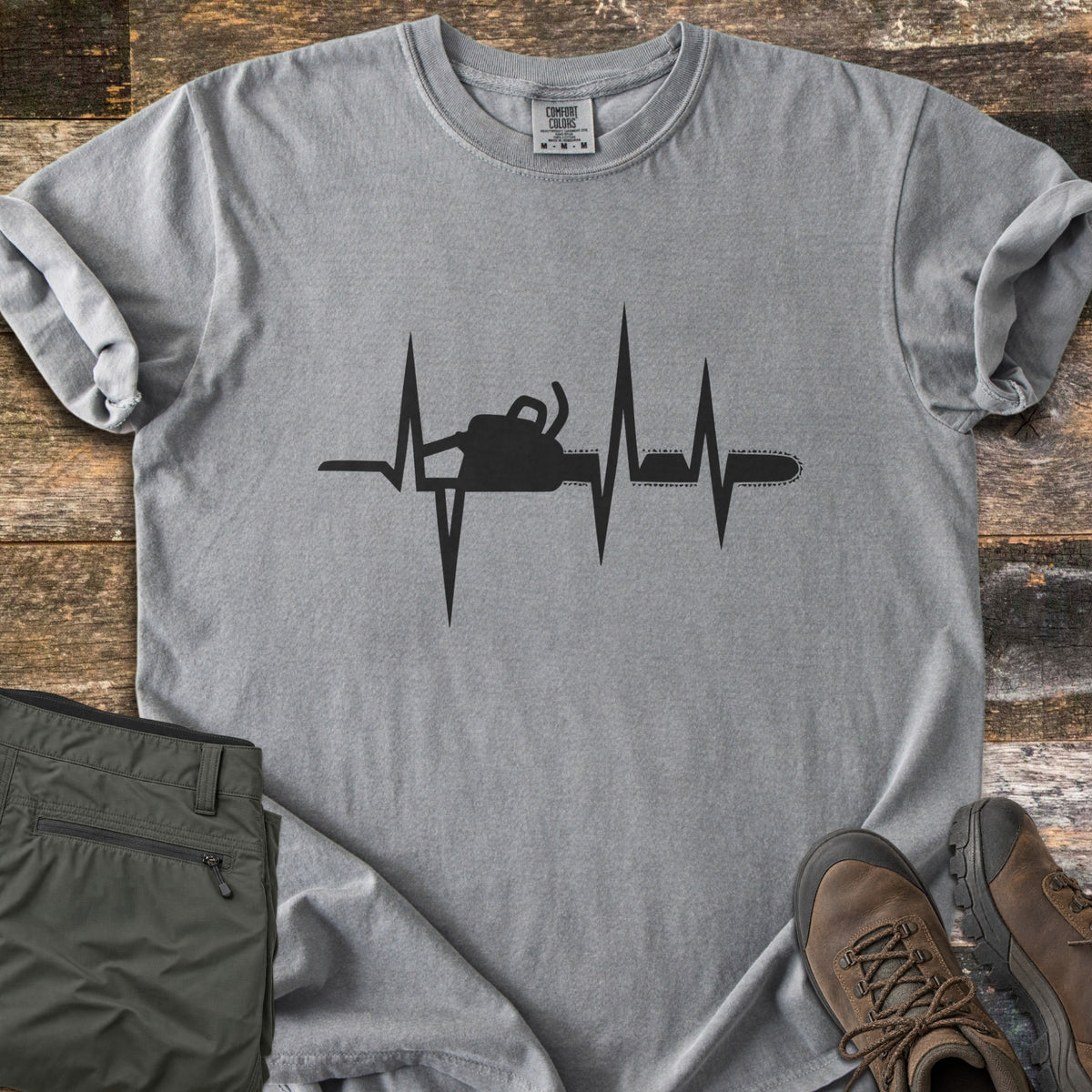 Chainsaw Heartbeat Comfort Colors T-shirt in Grey, laid flat. A black heartbeat line runs across the chest with a chainsaw silhouette built into it, the bar and chain extending to the right. Folded olive cargo shorts and brown hiking boots sit on a weathered wood floor.