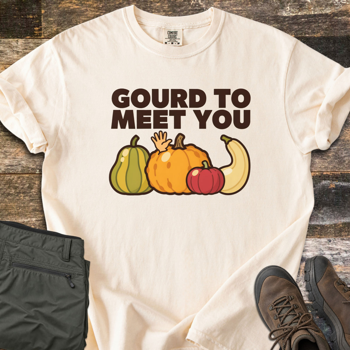 Gourd To Meet You Comfort Colors T-shirt in Ivory, laid flat. Dark brown words Gourd To Meet You above cartoon gourds: a green striped squash, an orange pumpkin with a waving hand, a red pumpkin and a cream squash. Olive pants and brown hiking boots sit on wood planks.