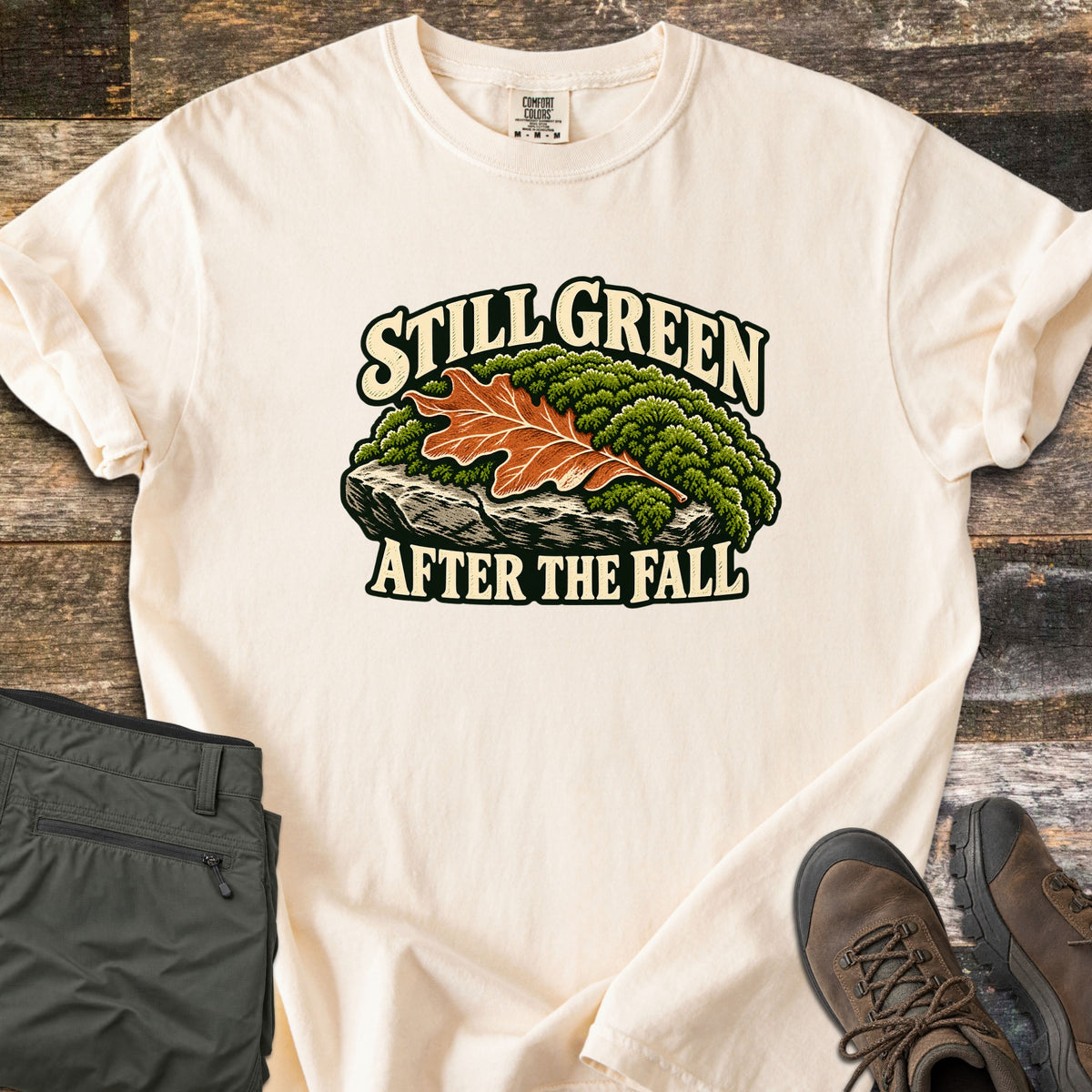 Still Green After the Fall Comfort Colors T-shirt in Ivory, laid flat. An orange oak leaf rests on green moss over a gray rock, with Still Green above and After the Fall below in cream letters outlined in dark green. Olive hiking pants and brown boots lie beside it on wood planks.