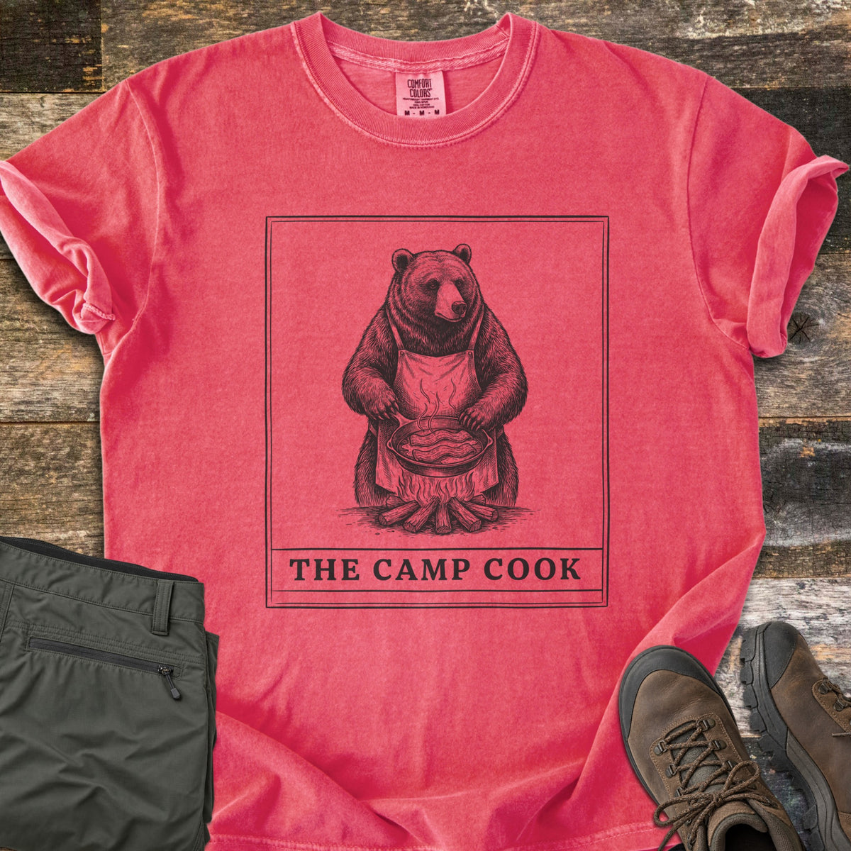The Camp Cook Comfort Colors T-shirt in Watermelon, laid flat. A framed sketch of a bear in an apron frying bacon in a skillet over a campfire, above the words THE CAMP COOK. Sleeves are cuffed on weathered wood beside olive hiking pants and brown hiking boots.