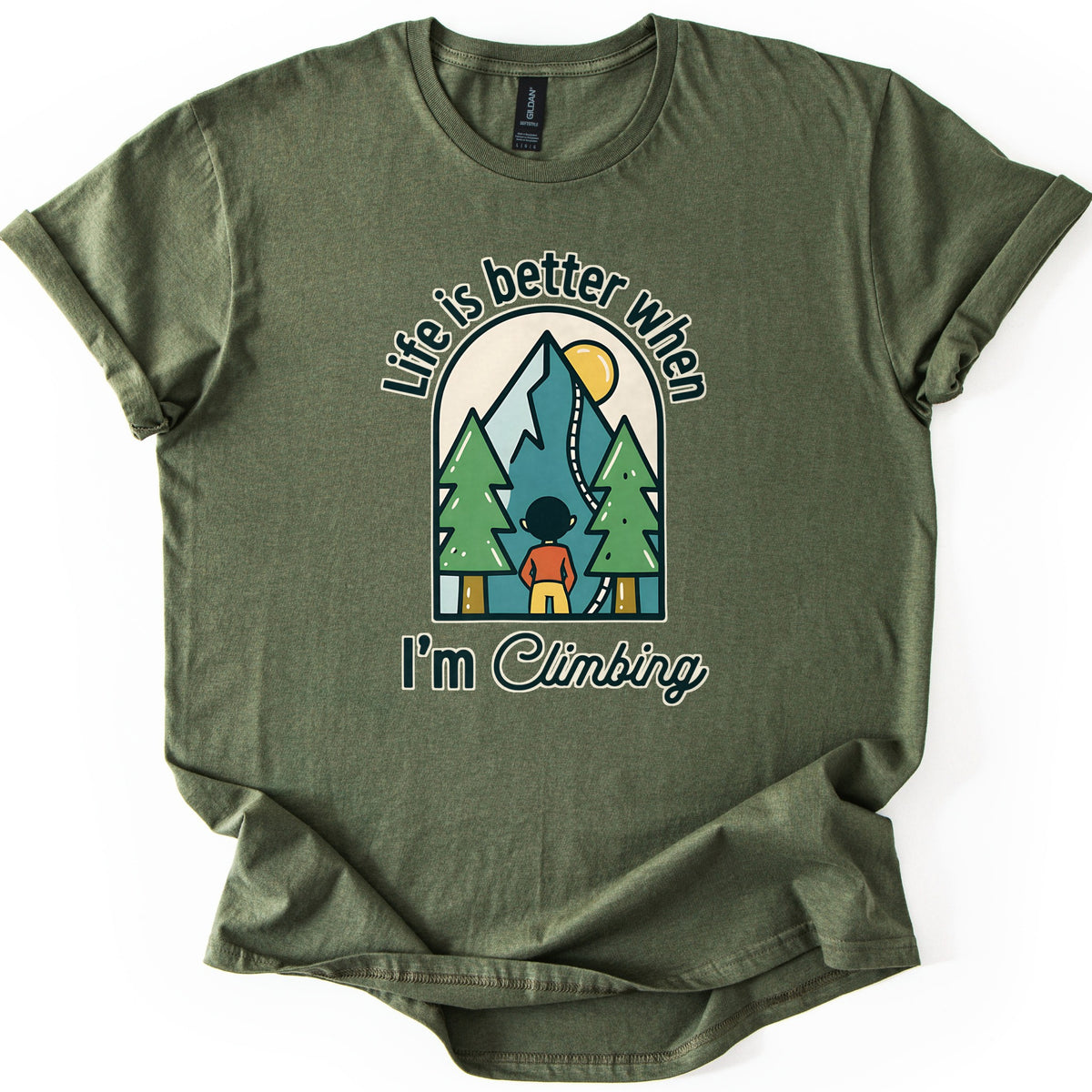 Better When I'm Climbing Comfort Colors T-shirt in Military Green, laid flat. The words Life is better when arch over a window scene of a person facing a teal mountain with a winding trail, a yellow sun and two pine trees, with I'm Climbing below. Sleeves cuffed, on a white surface.