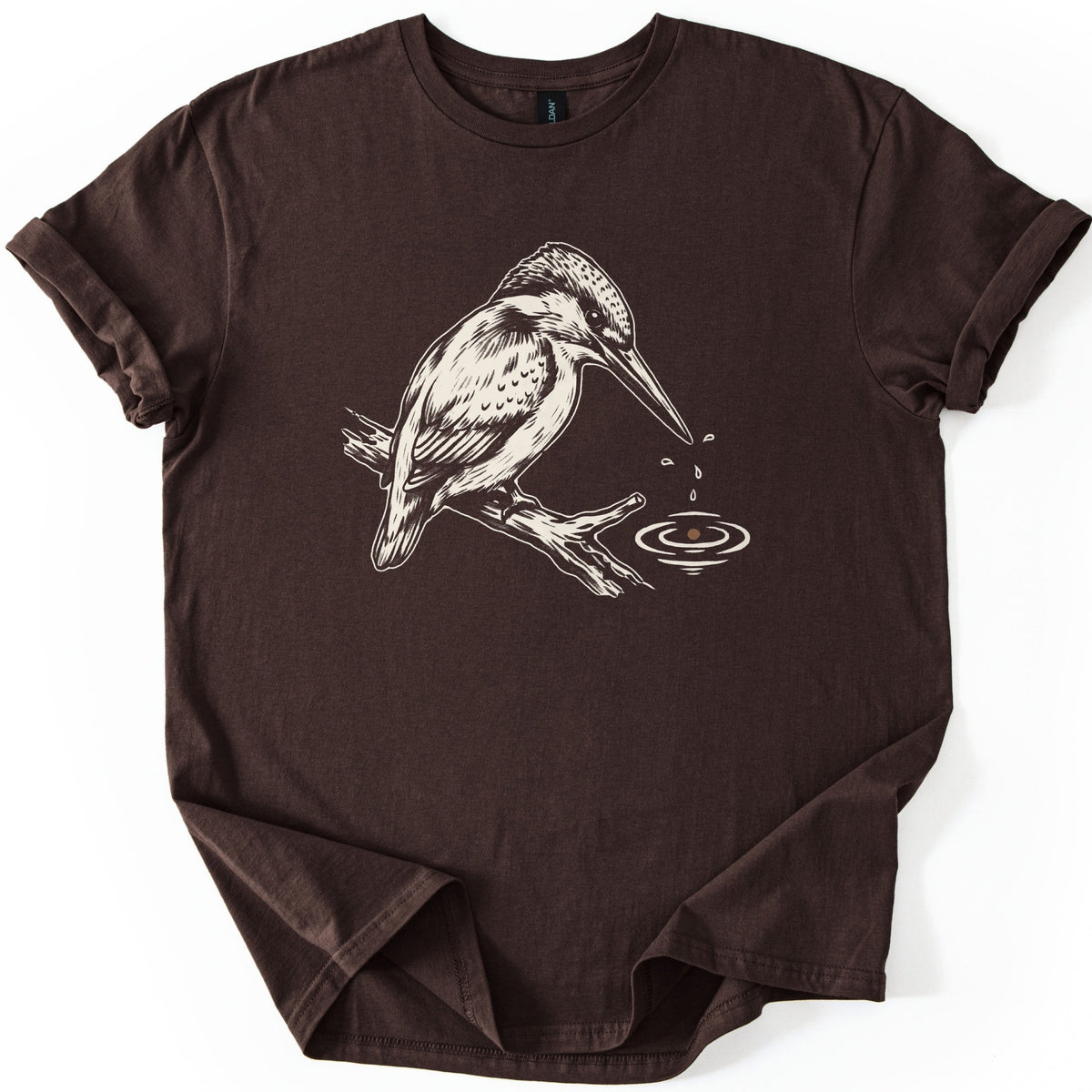 Kingfisher Comfort Colors T-shirt in Dark Chocolate, laid flat. A cream-colored drawing of a kingfisher perched on a branch, with droplets falling from its beak into rippling water below. The sleeves are cuffed and the shirt lies on a white background.