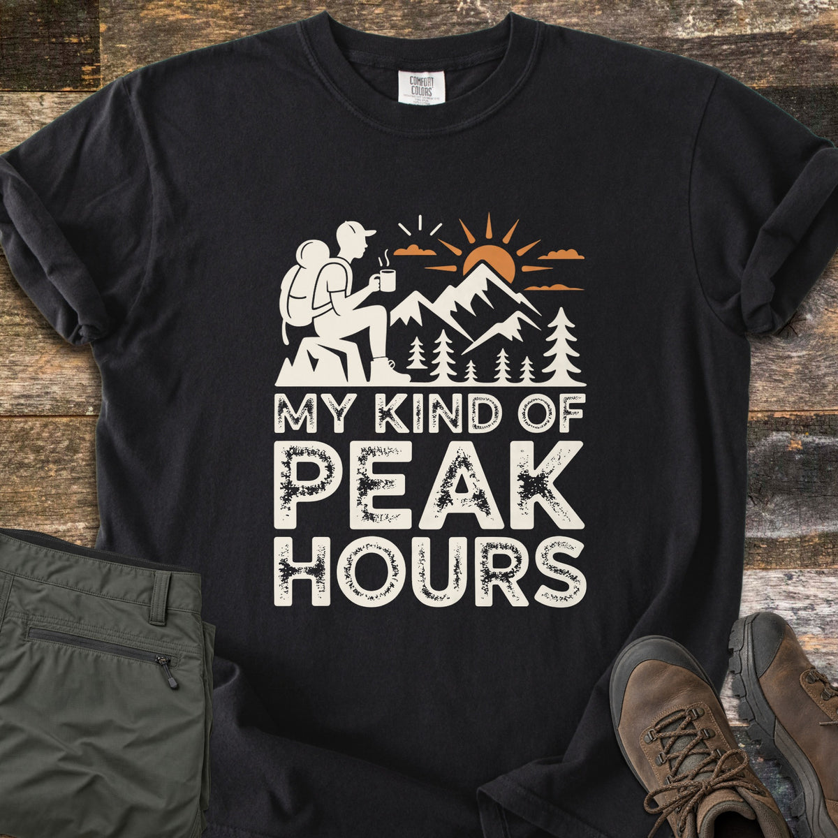 My Kind Of Peak Hours Comfort Colors T-shirt in Black, laid flat. A cream hiker with a backpack sits on a rock with a steaming mug, facing mountains, pines and an orange sun, above the words My Kind Of Peak Hours. Olive hiking pants and brown boots lie on weathered wood.