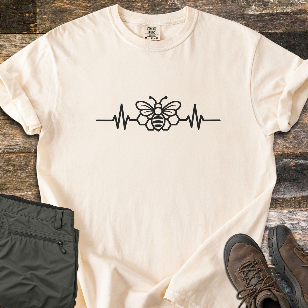 Bee Heartbeat Comfort Colors T-shirt in Ivory, laid flat. A black line drawing of a bee over honeycomb hexagons sits in the middle of a heartbeat line across the chest. Sleeves are rolled, on weathered wood beside olive hiking pants and brown hiking boots.