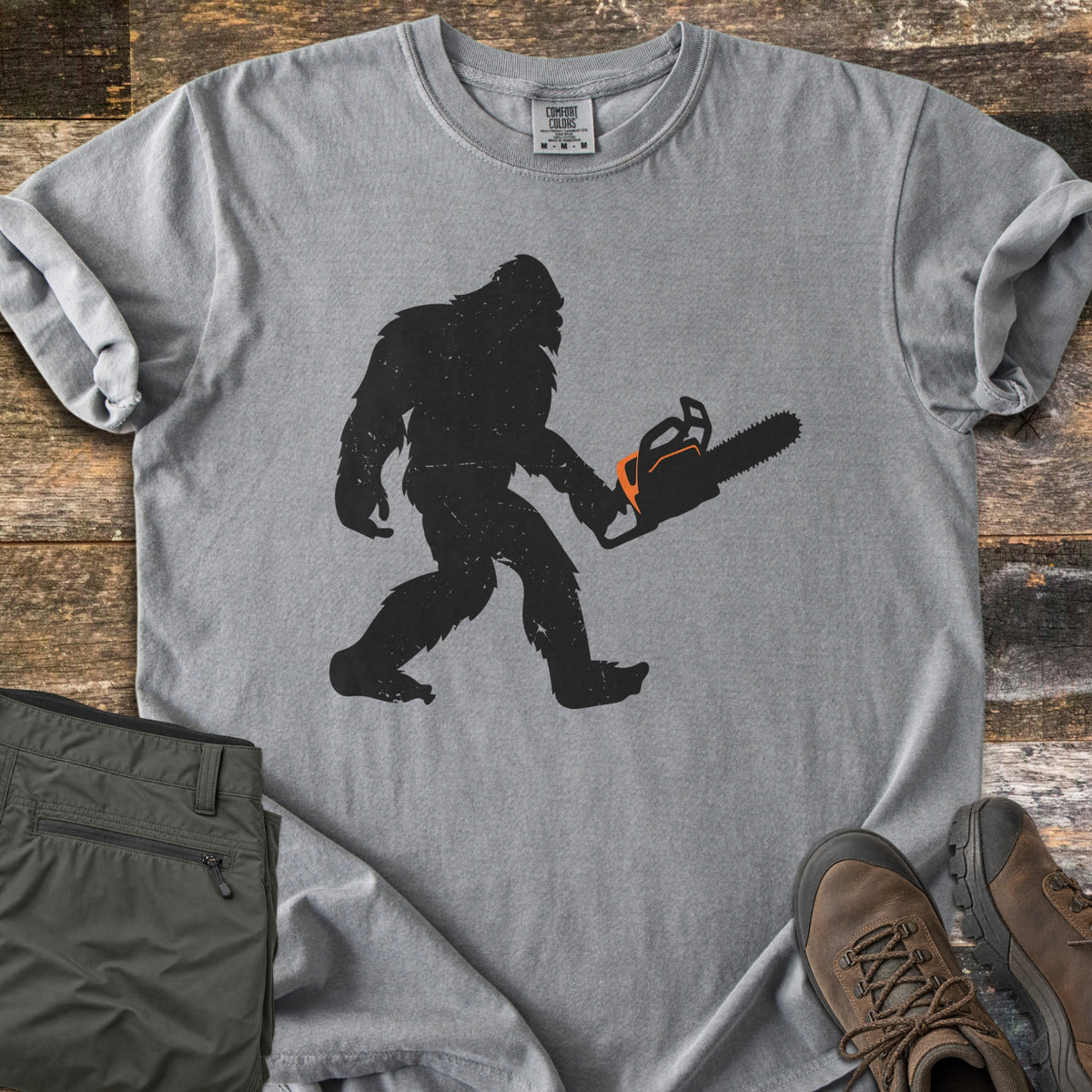 Lumber Squatch Comfort Colors T-shirt in Grey, laid flat. A distressed black silhouette of Bigfoot walks while carrying a chainsaw with orange accents. The sleeves are rolled, with folded olive pants and brown hiking boots on rustic wood planks.